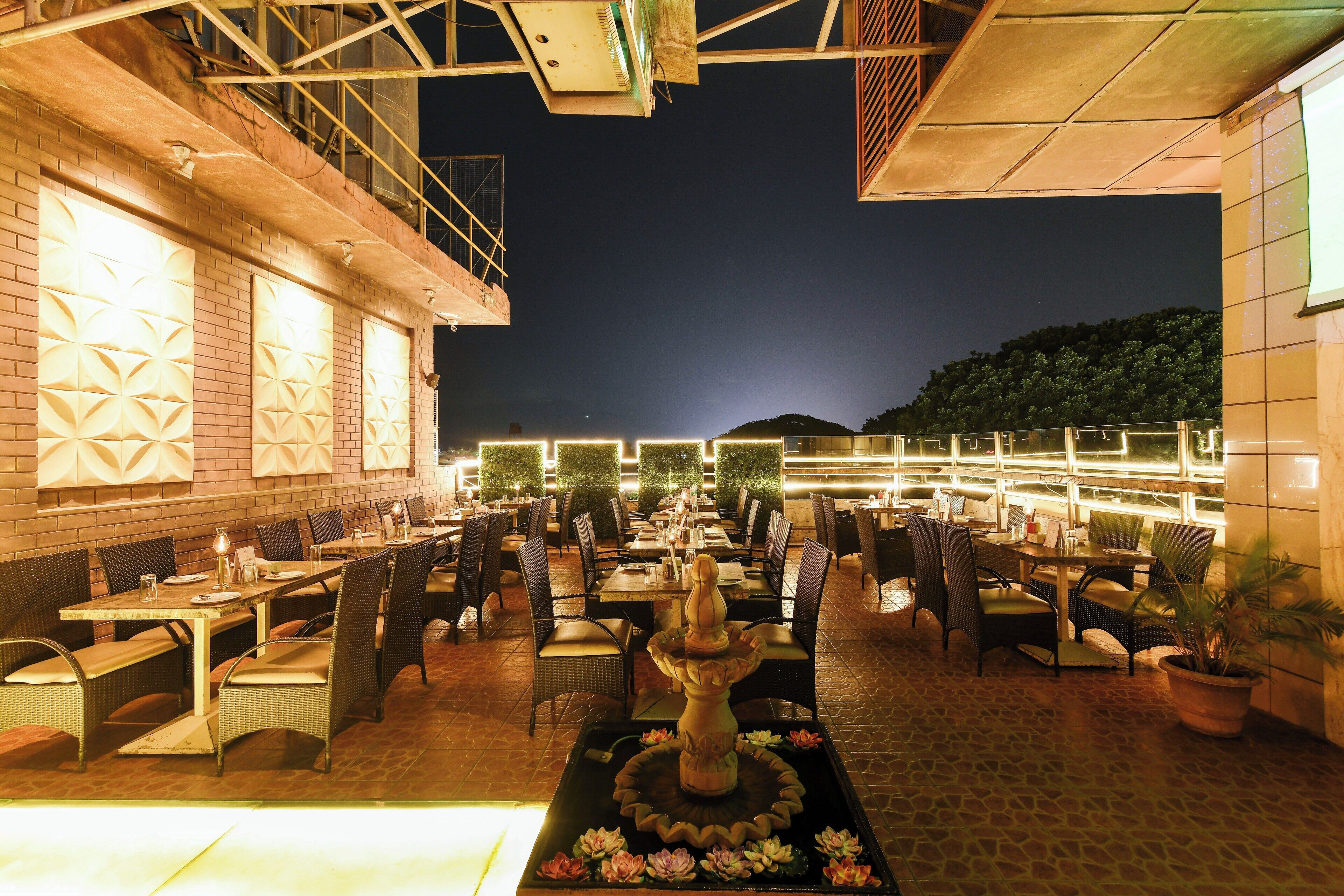 An outdoor dining area with brick walls, multiple tables and chairs, and a central decorative fountain.