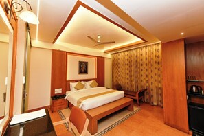 A hotel room with a large bed, a desk, a chair, a ceiling fan, a window with curtains, and a kitchenette.