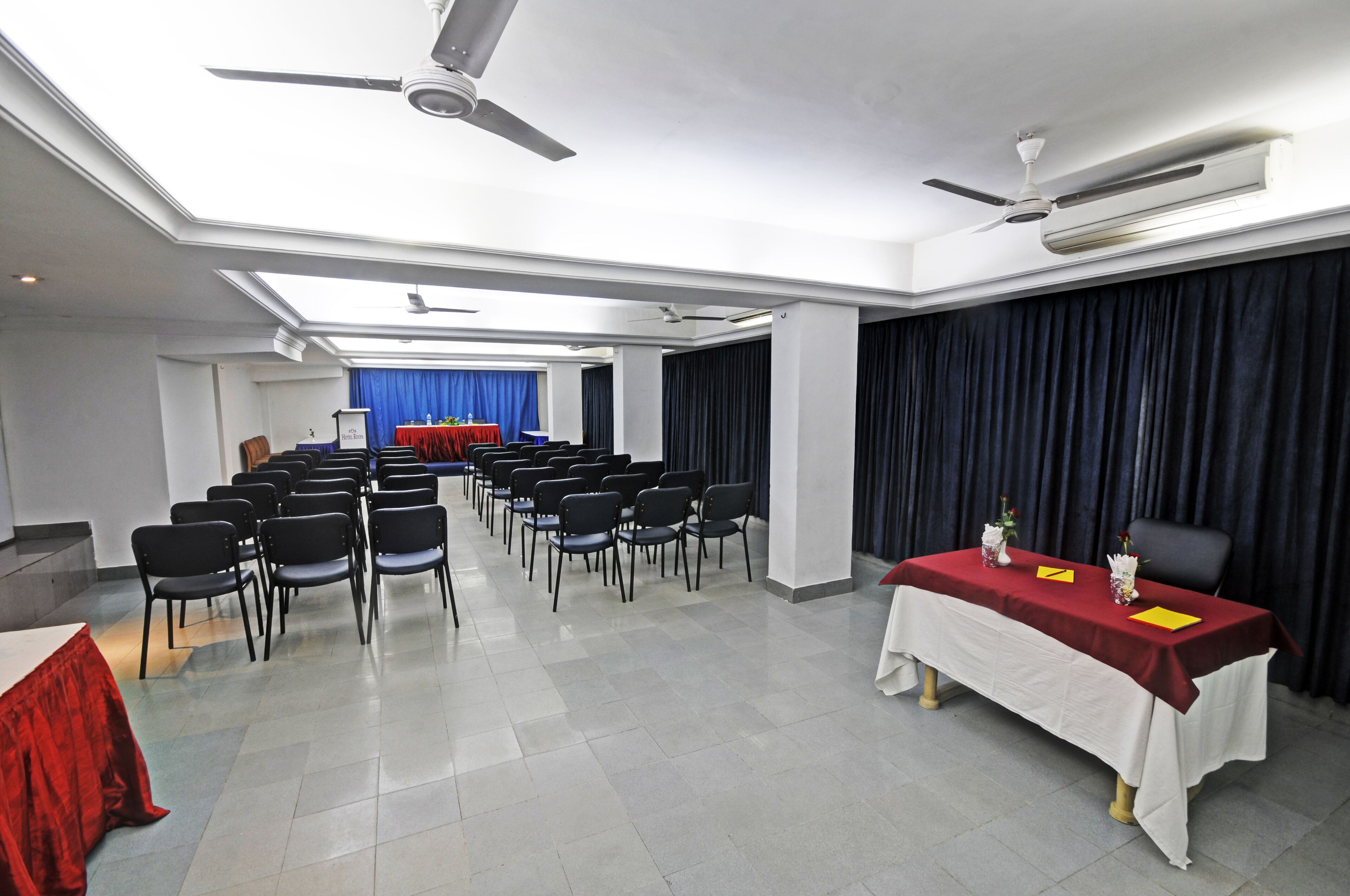 A conference room with rows of chairs, a table with a red tablecloth, and a ceiling fan.