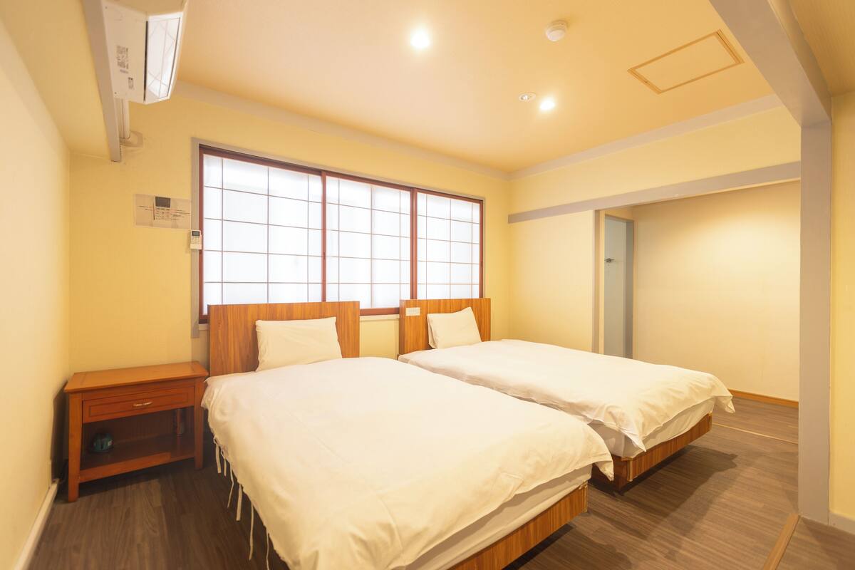 Habitación de hotel con dos camas, una mesita de noche de madera, un ventanal con diseño de cuadrícula y una lámpara en el techo.
