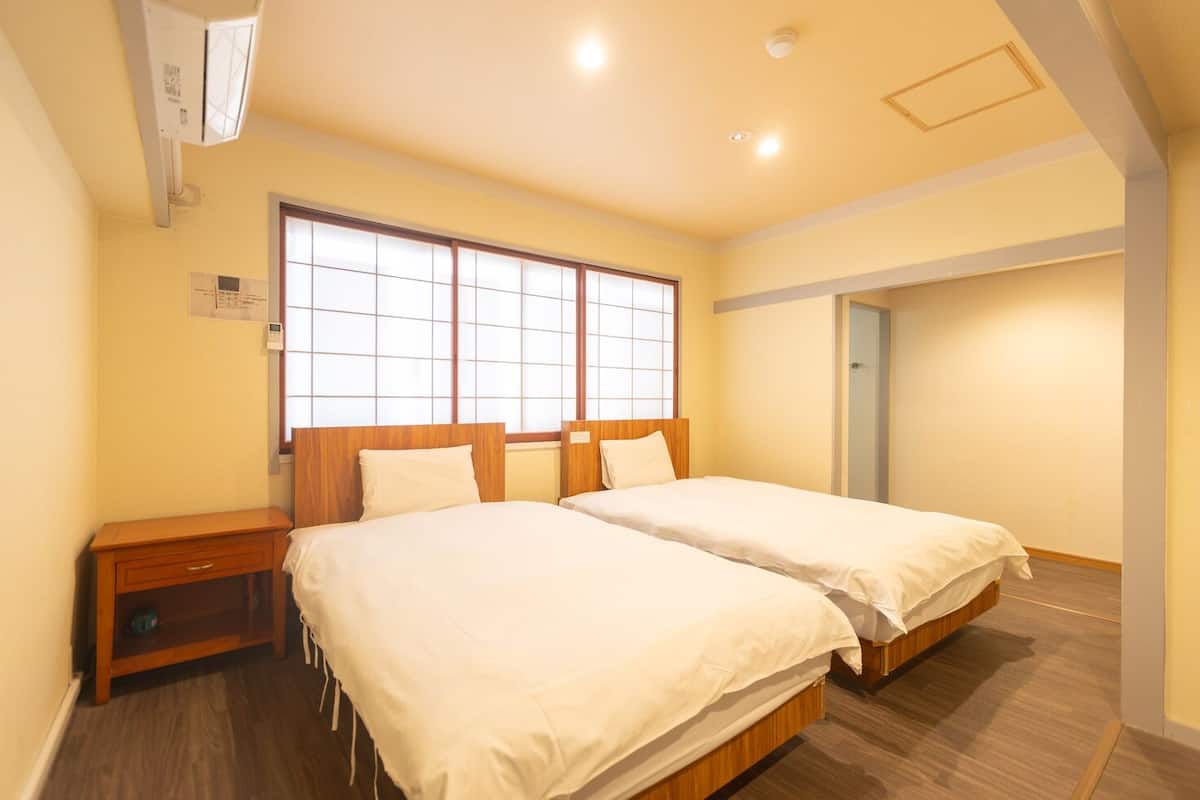 Une chambre d’hôtel avec deux lits, une table de chevet en bois, une fenêtre avec des stores et une porte.