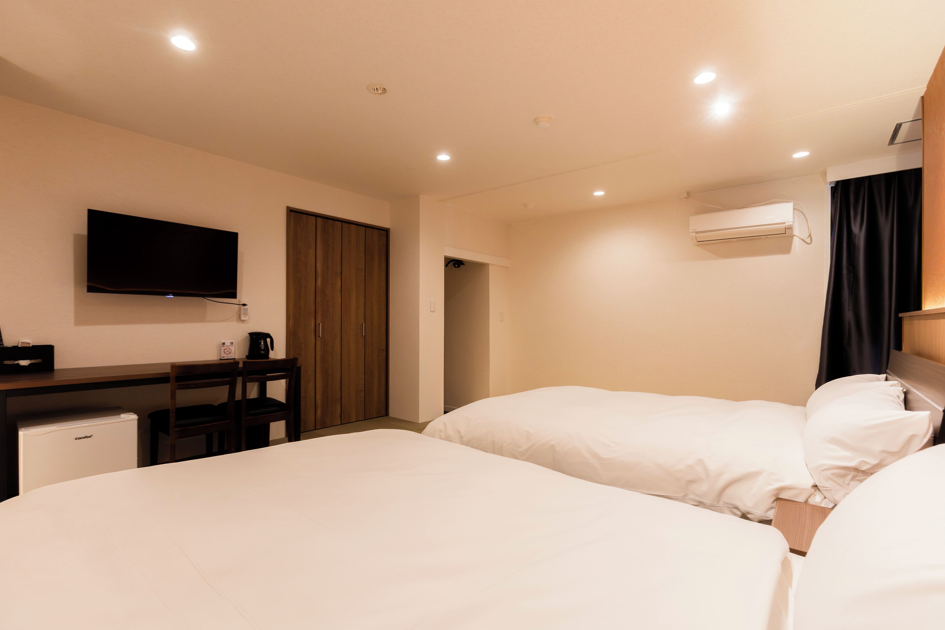 A hotel room with two beds, a desk, a television, and an air conditioning unit.