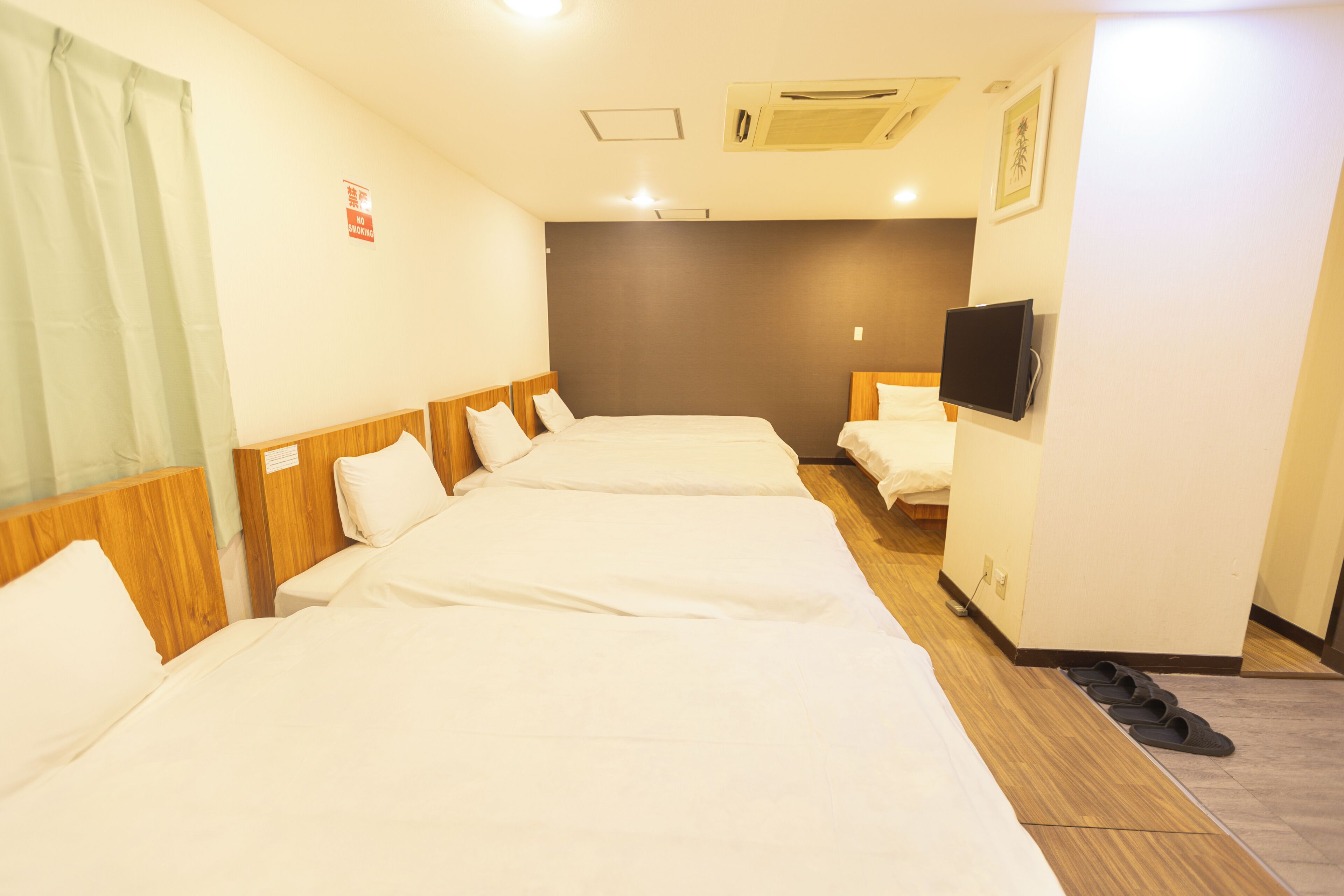 A hotel room with three single beds, a television mounted on the wall, and a pair of slippers on the floor.