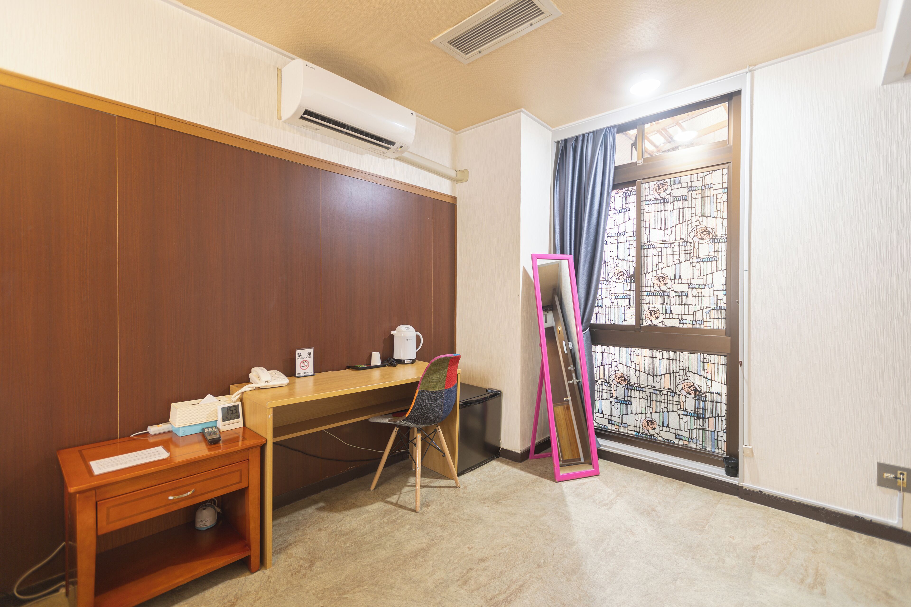 Habitación de hotel con escritorio de madera, una silla, una mesita con teléfono y reloj, y ventana con un diseño de vitral.