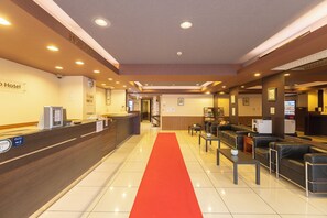 Zona de recepción de un hotel con alfombra roja, mostrador de recepción, área de estar y un reloj en la pared.
