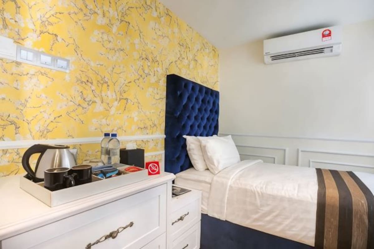 A hotel room with a bed, a dresser, a nightstand, a wall-mounted air conditioner, and a floral wallpaper.