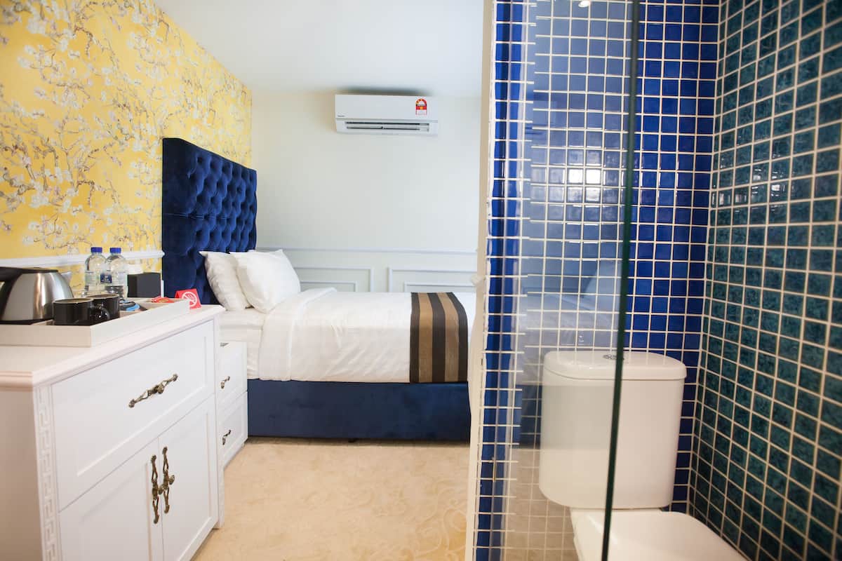 Una camera d'albergo con un letto, una cassettiera bianca, un condizionatore d'aria e un bagno con piastrelle azzurre.