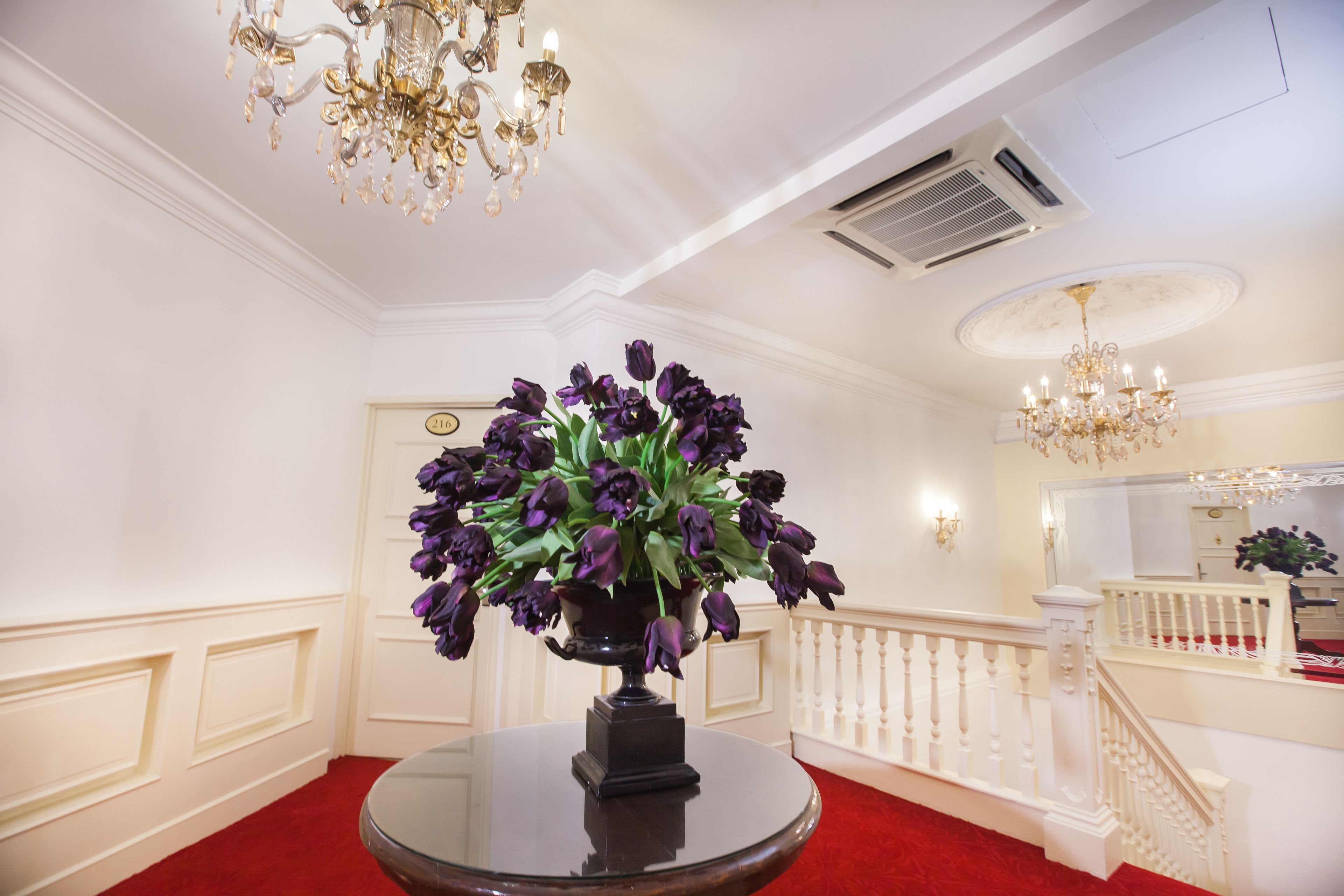 Un amplio pasillo con una mesa redonda adornada con un jarrón de flores moradas, un candelabro y una escalera con barandilla blanca.