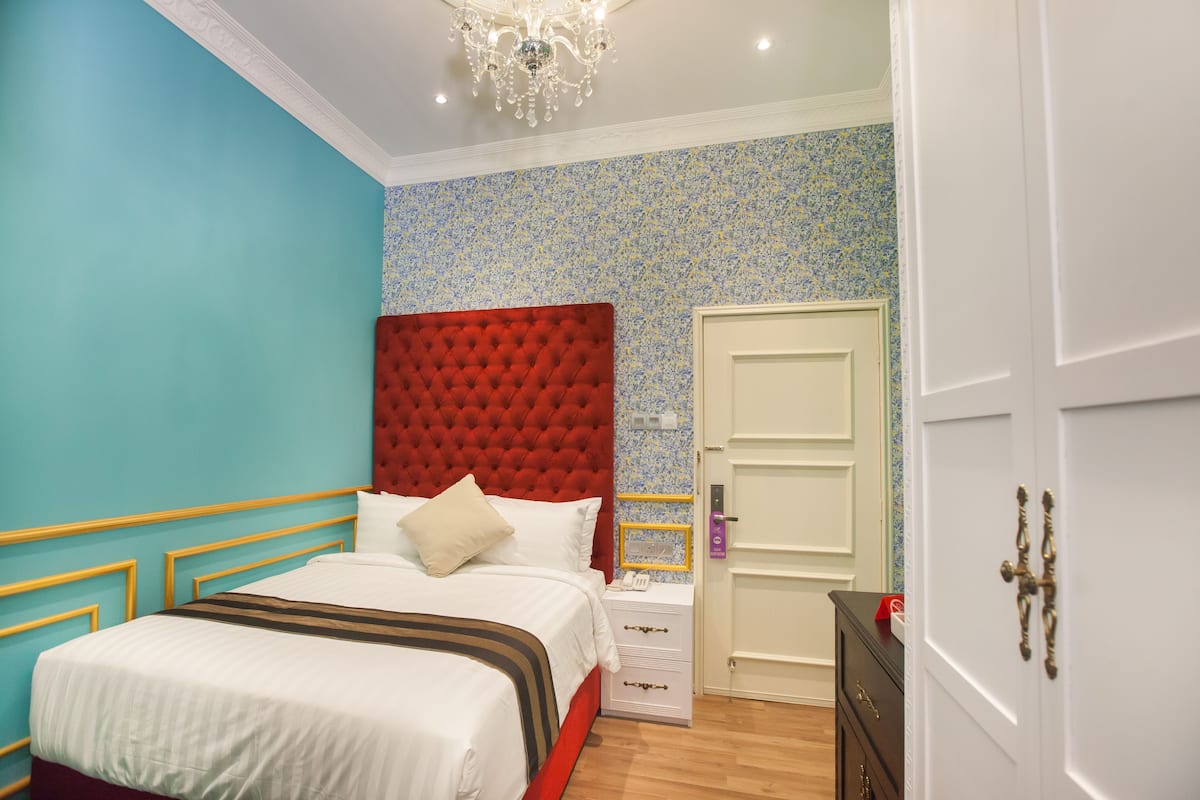 Una camera da letto con una testiera imbottita rossa, un letto bianco con coperta a righe, un comodino con telefono e specchio, e una porta con maniglia in ottone.