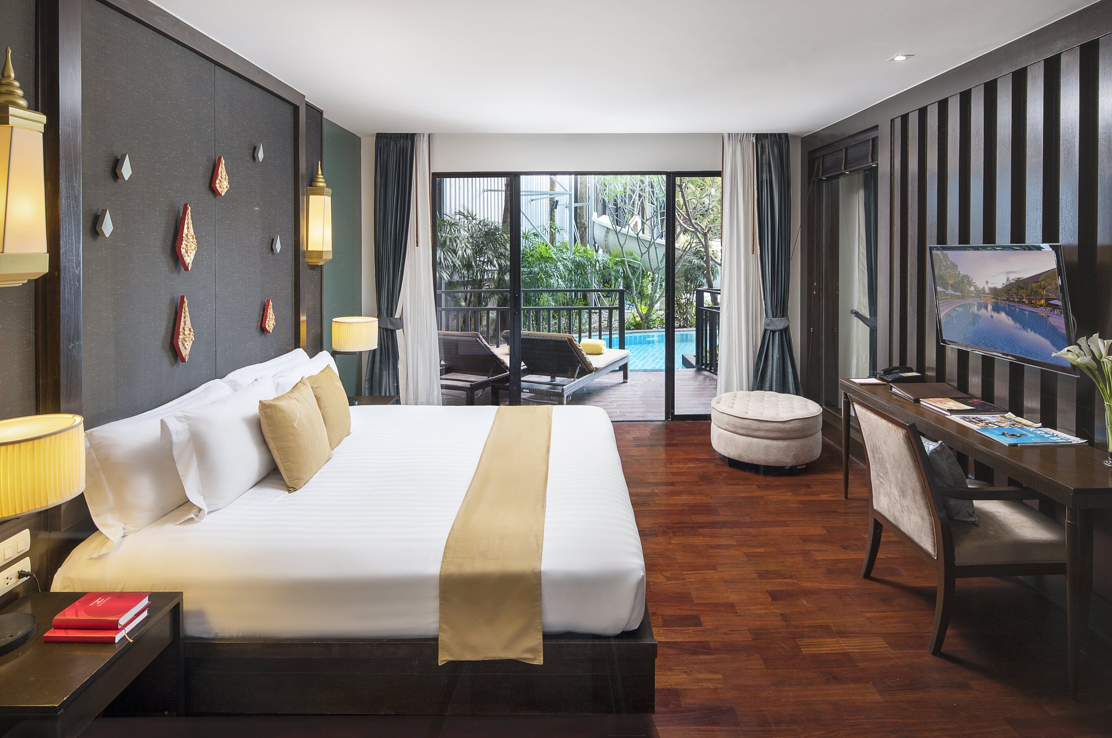 A hotel room with a large bed, a desk with a chair, a TV, and a balcony with outdoor seating.