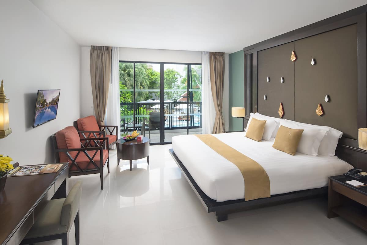 Une chambre d’hôtel avec un grand lit, un bureau avec une chaise, une petite table avec une lampe, et une vue sur une piscine et la verdure par la fenêtre.