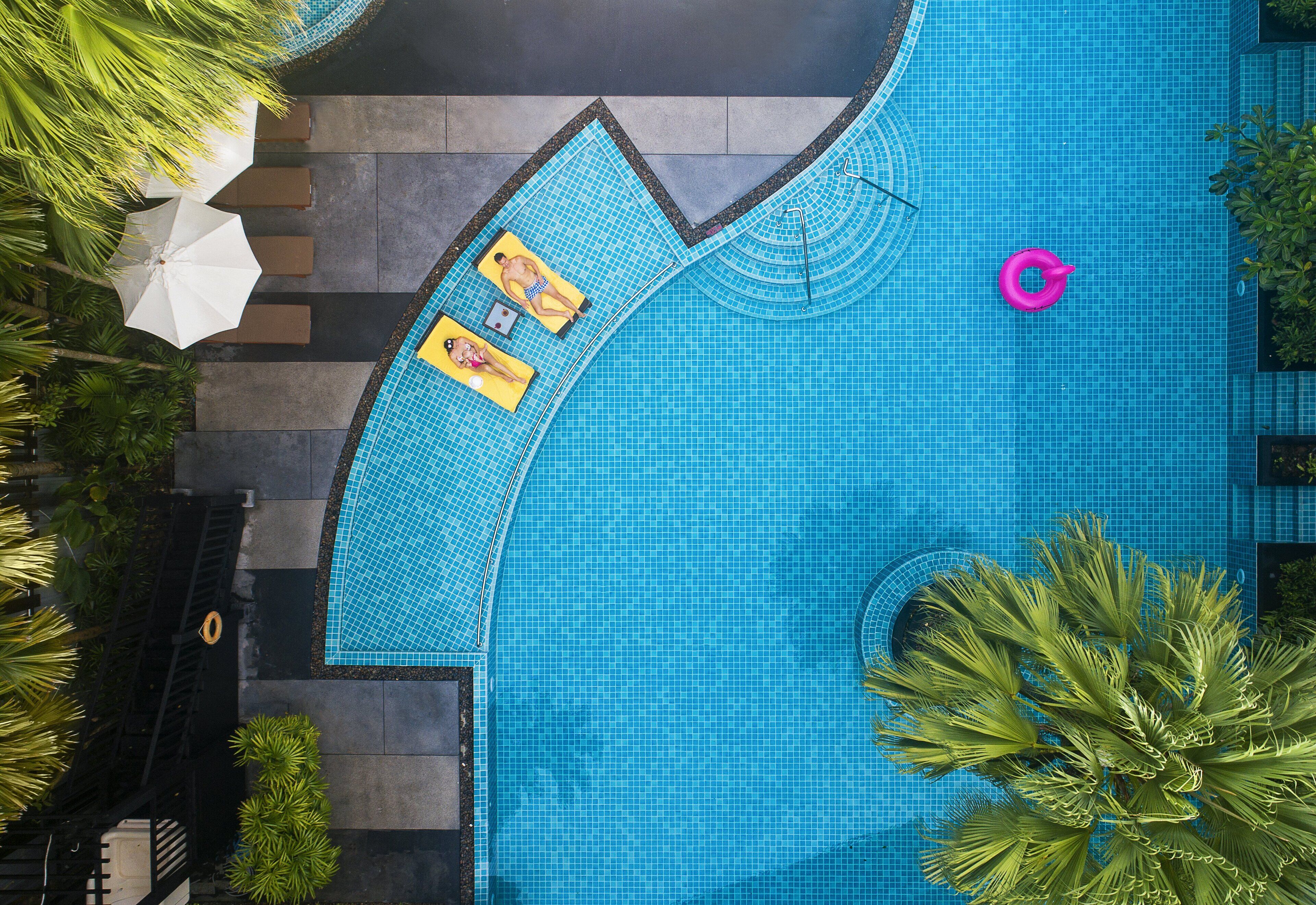 A large swimming pool with a tiled edge, surrounded by palm trees and a few umbrellas.