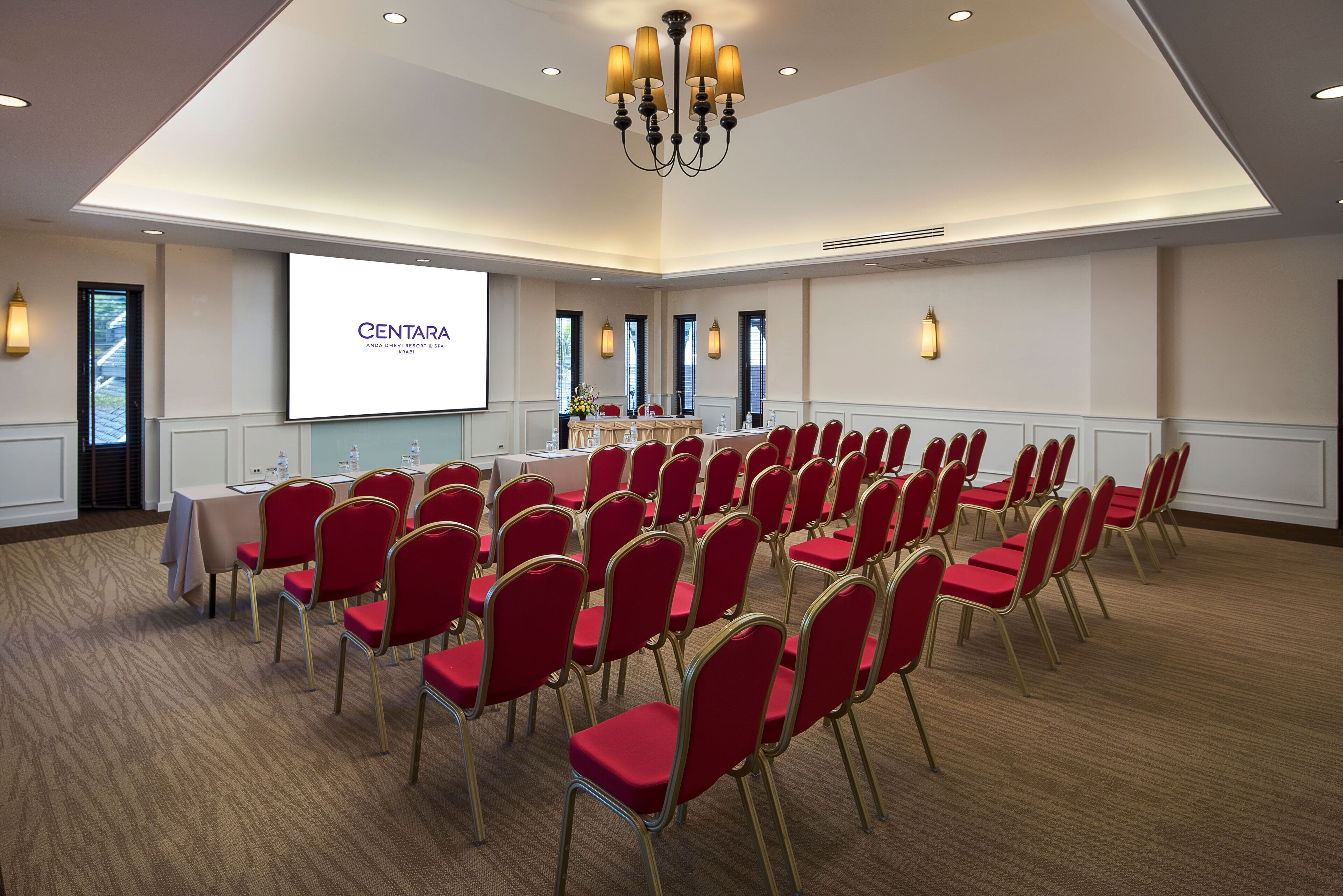 A conference room with rows of red chairs, a projector screen displaying 'CENTARA ANDA DHEVI RESORT & SPA KRABI', and a chandelier.