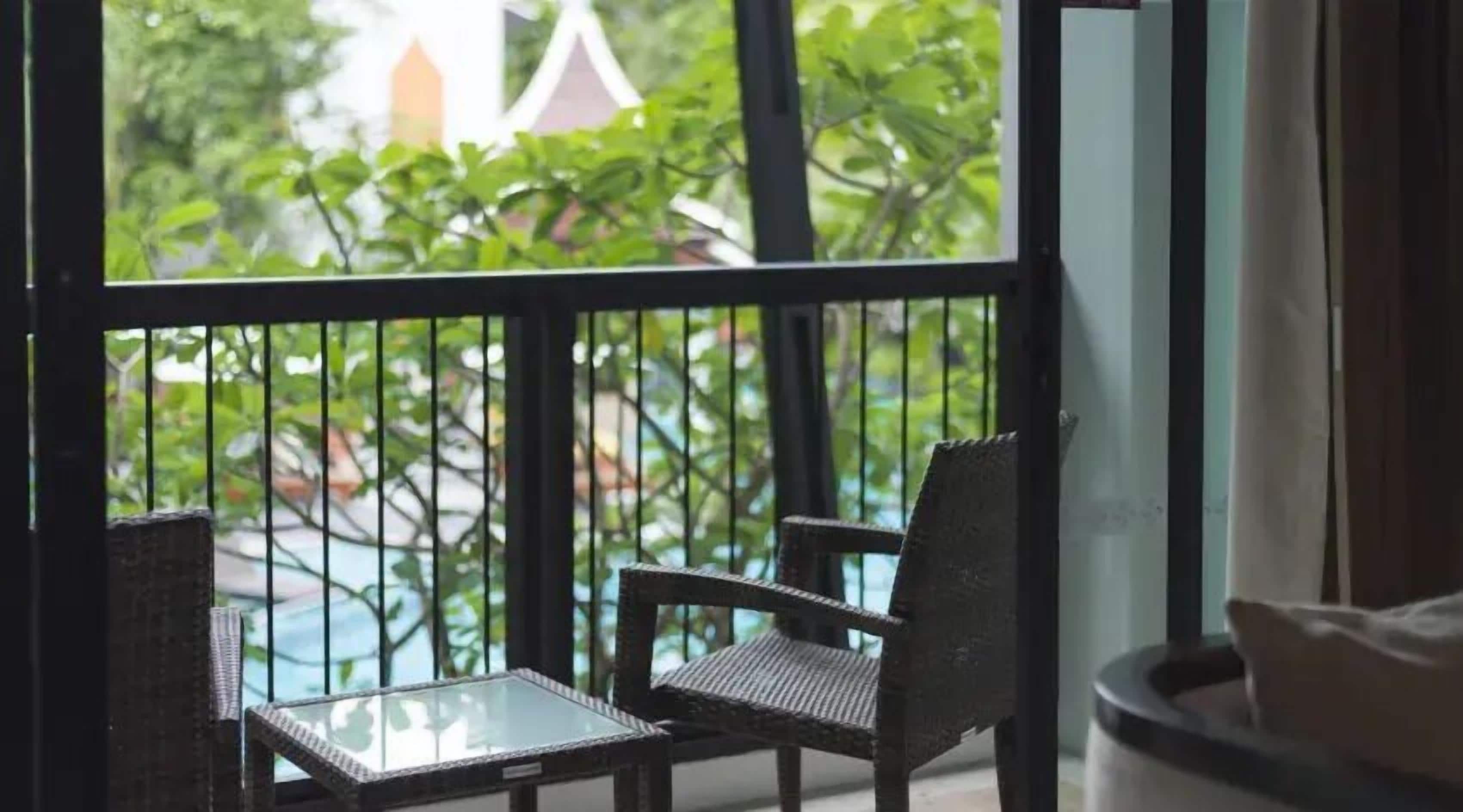 A balcony with two wicker chairs and a small glass table, overlooking a pool and greenery.