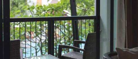A balcony with two wicker chairs and a small glass table, overlooking a pool and greenery.