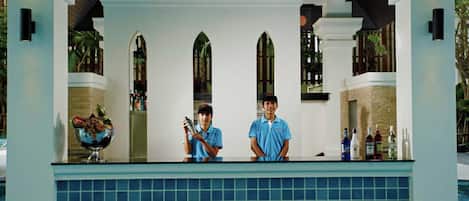 Un bar junto a la piscina con dos empleados, una piscina con baldosas y una pared de piedra.