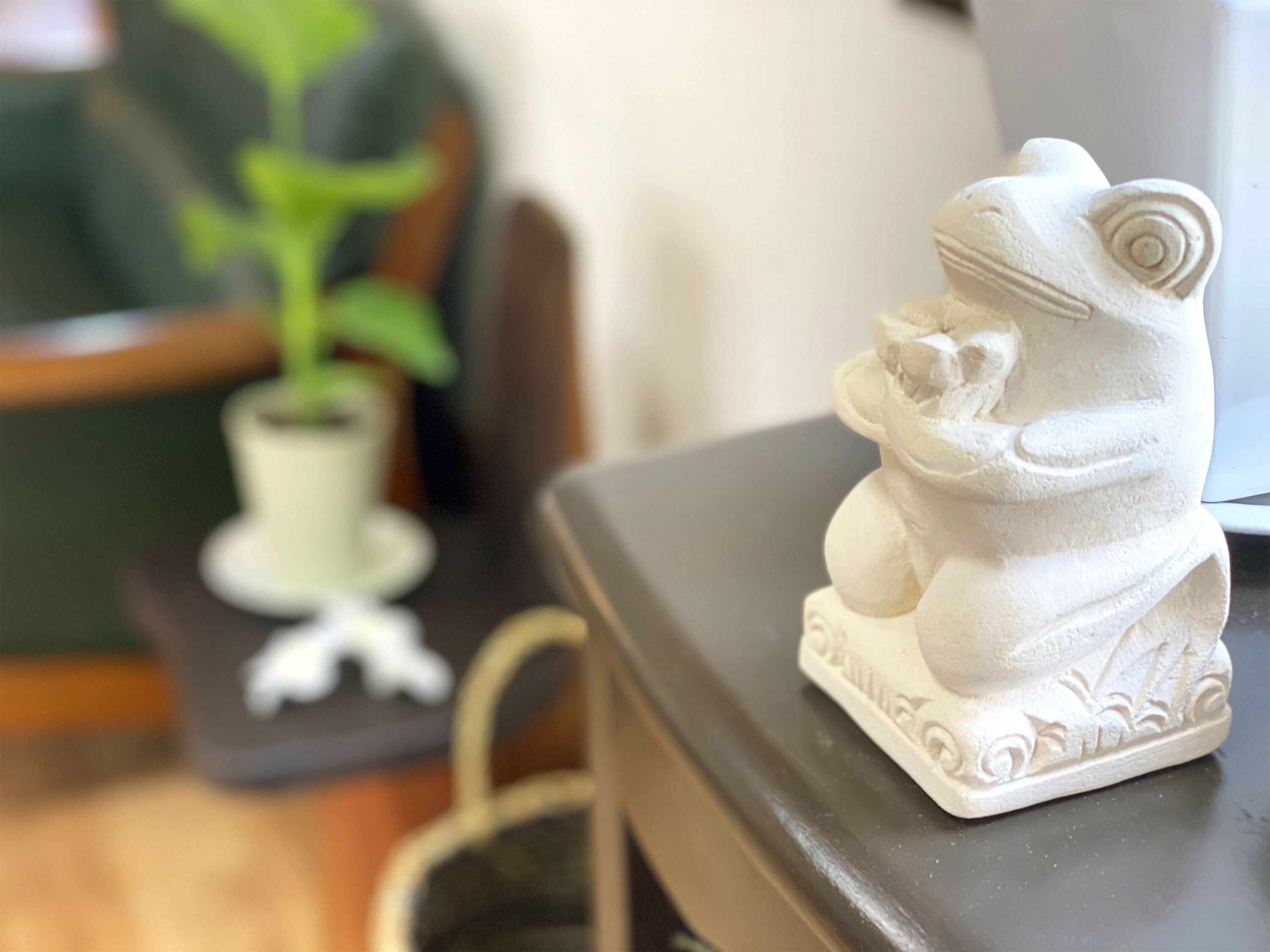 A white ceramic frog figurine on a dark surface, a small potted plant, and a chair.