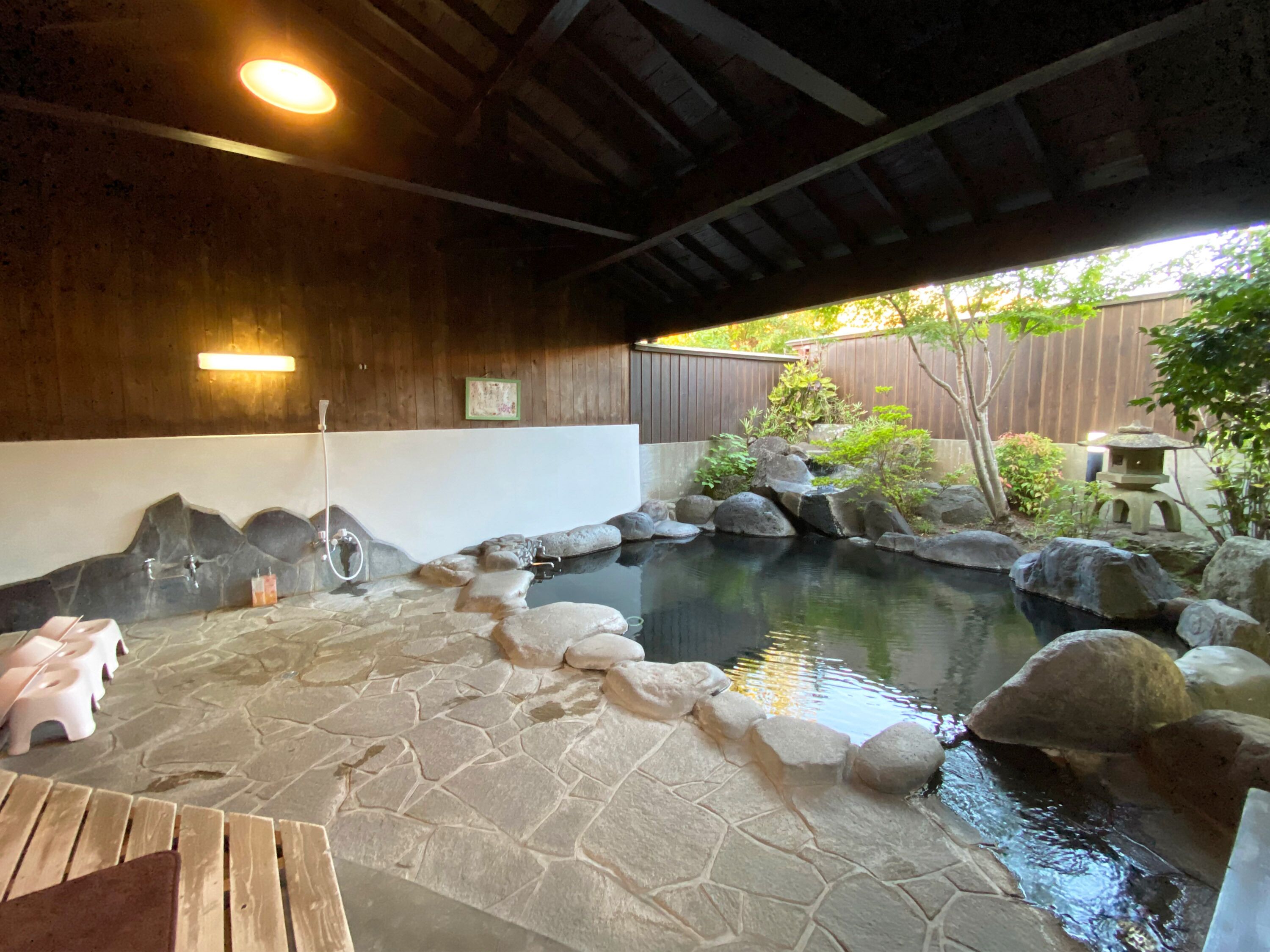 A stone-tiled hot tub area with a wooden ceiling, a small waterfall feature, and a wooden deck with pink stools.