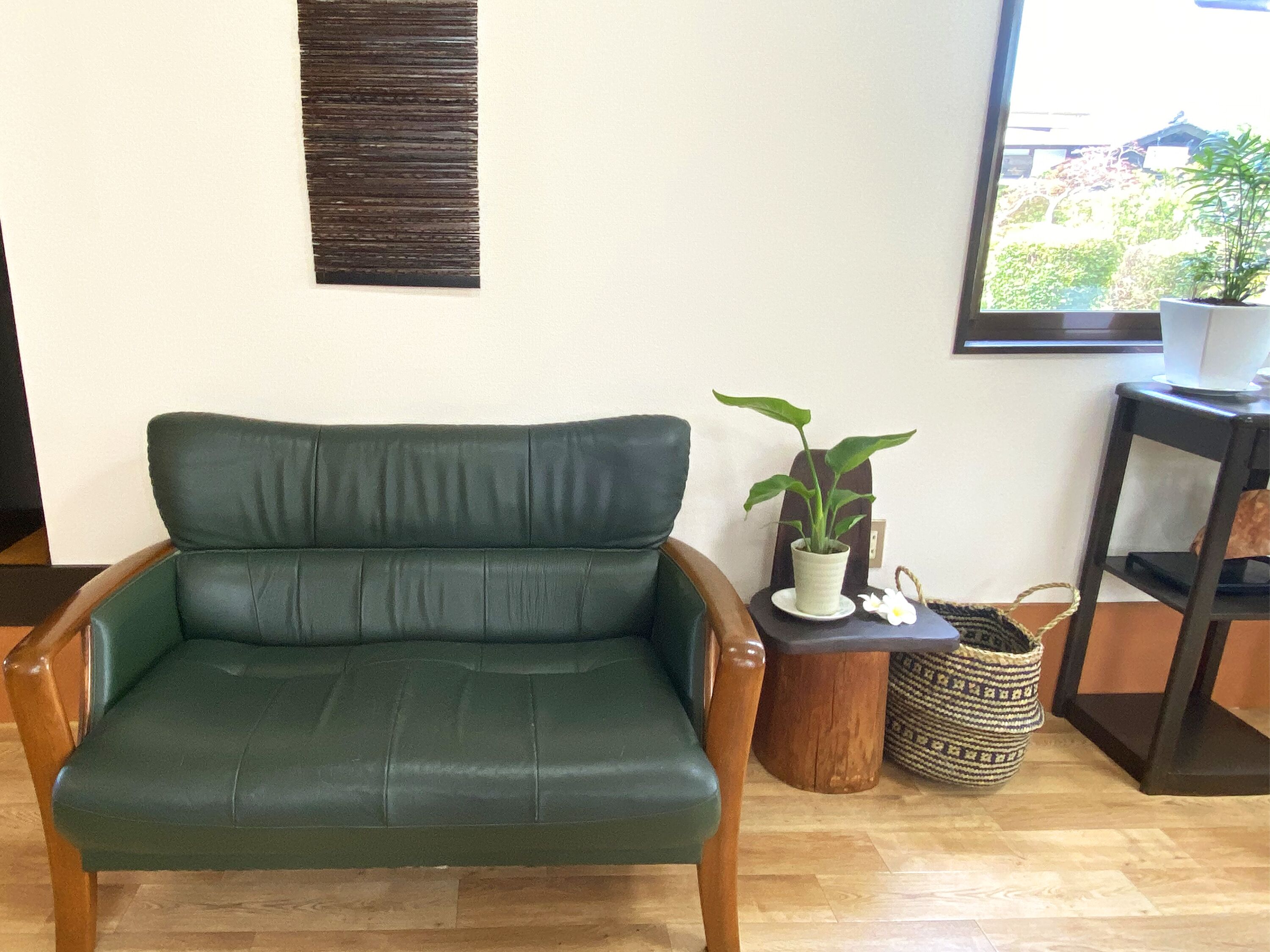 Un sillón de cuero verde en una habitación con suelo de madera, una pequeña mesa de madera con una planta en maceta, una cesta tejida y una ventana con vistas a la vegetación.