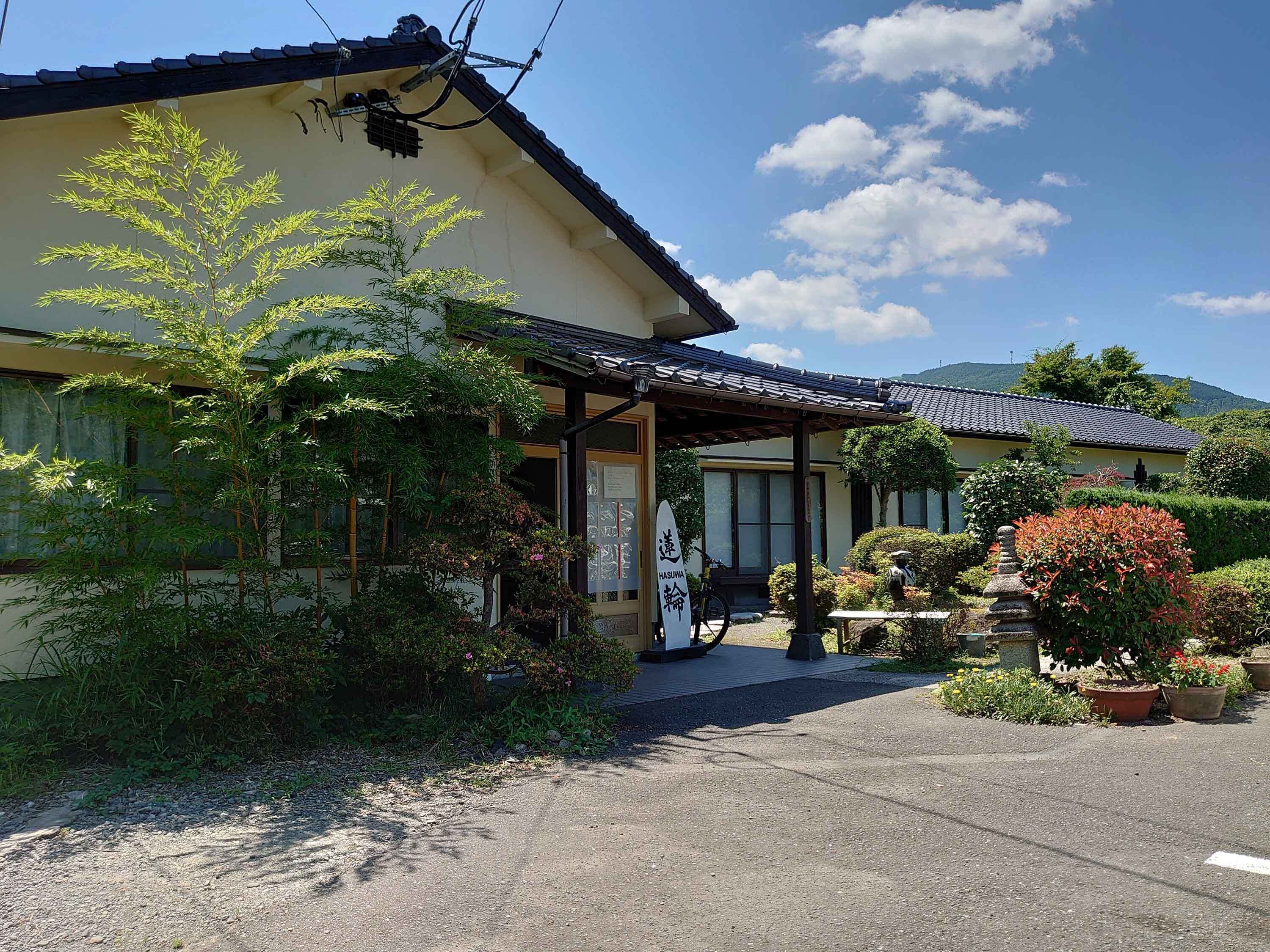 A traditional Japanese building with a tiled roof, a covered entrance, and a garden with various plants and a stone lantern.