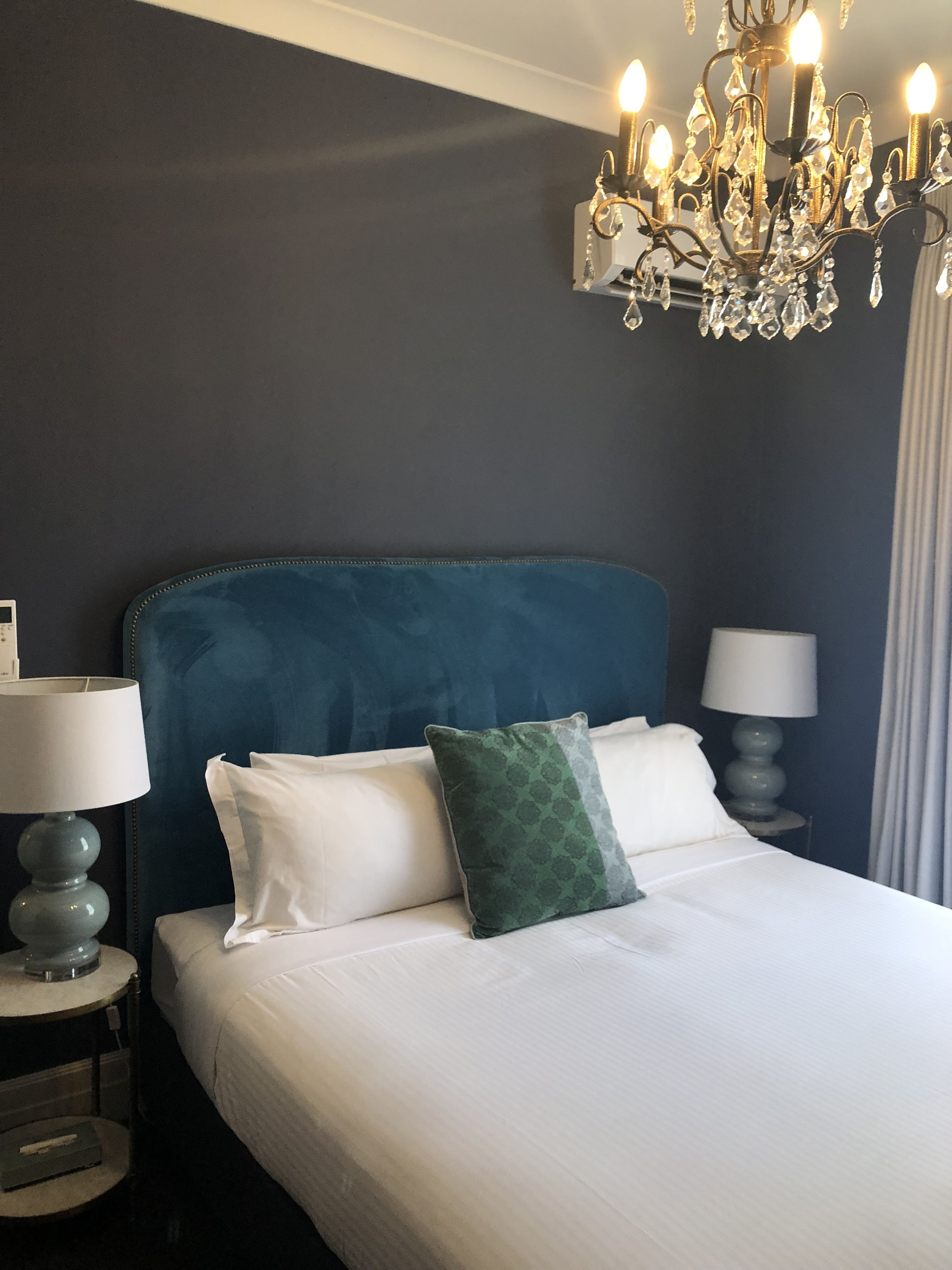 Une chambre avec un grand lit, deux tables de chevet, un lustre et un mur de couleur sombre.