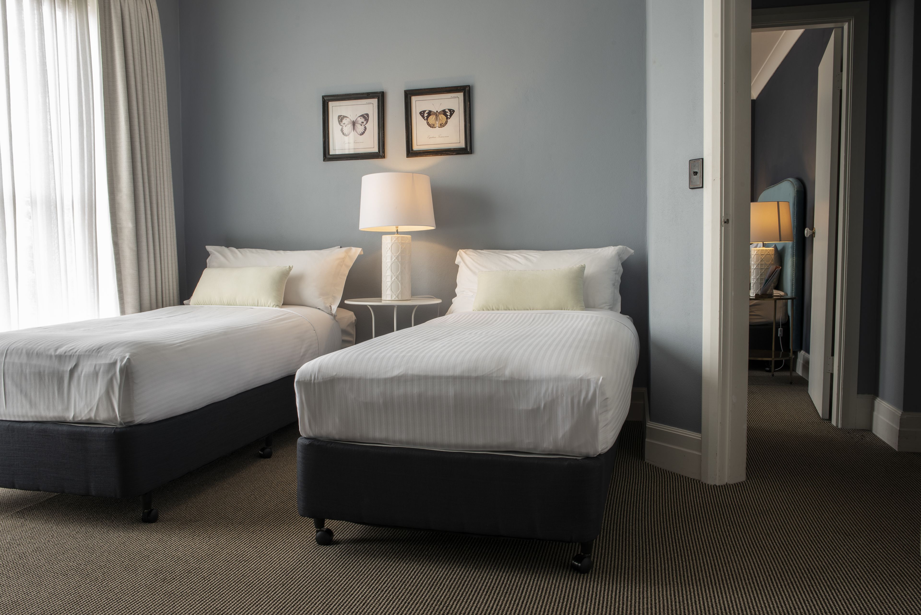 A hotel room with two beds, a nightstand with a lamp, and a door leading to another room.