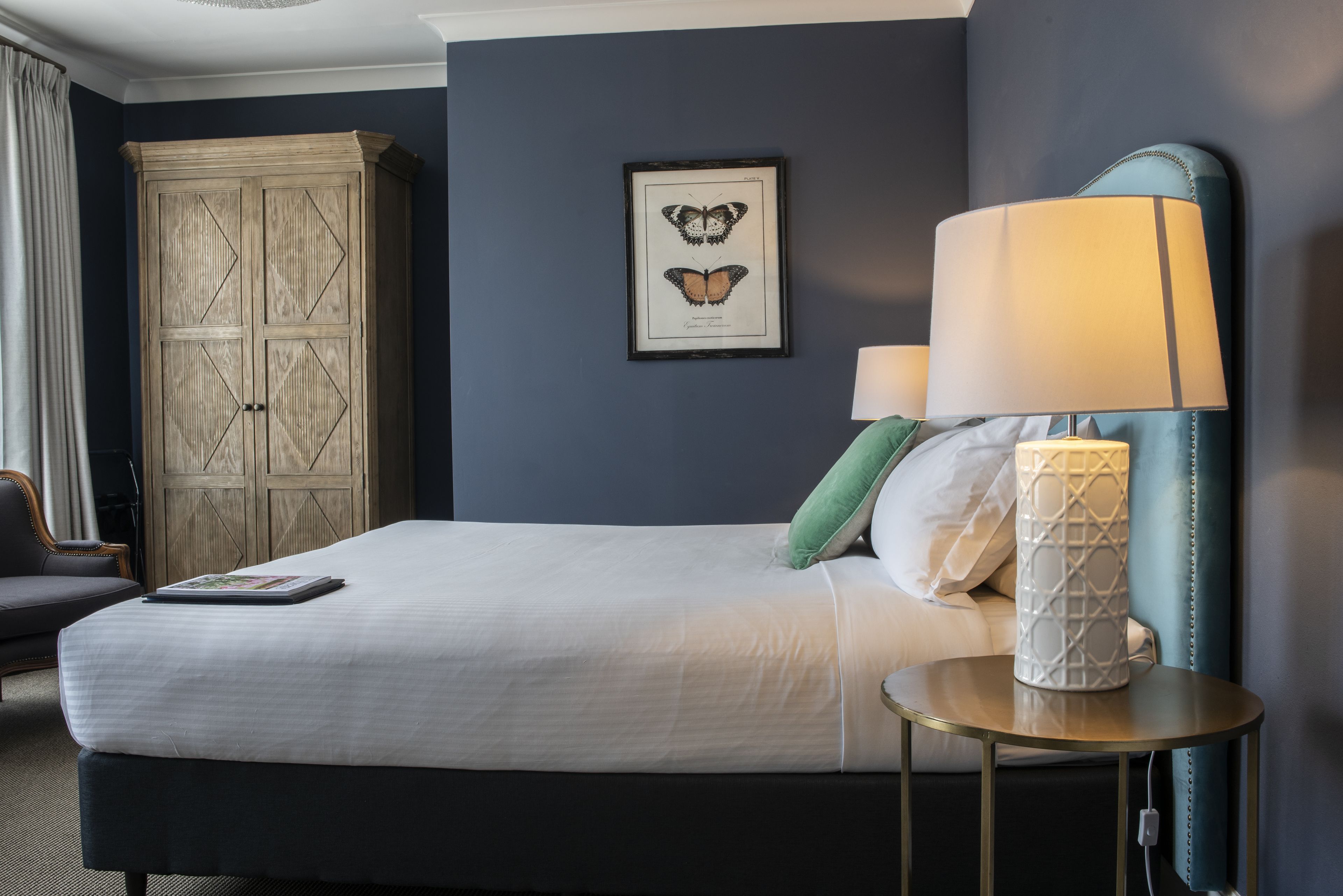 Een net opgemaakt bed met kussens, een bedlampje en een lijst met een vlinder afbeelding aan de muur.