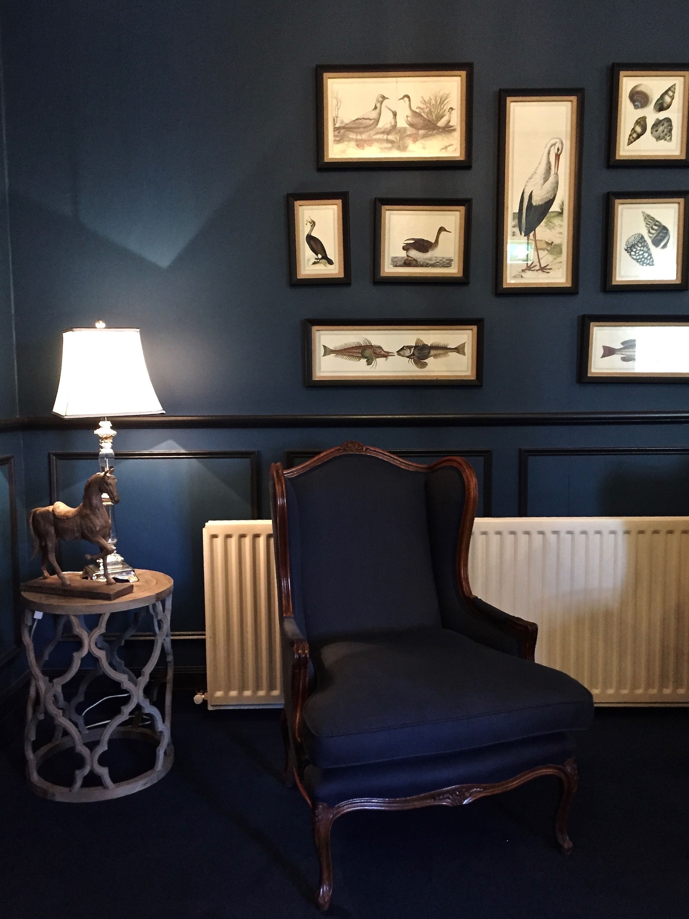 Una habitación con una pared azul oscuro, una silla de madera, una mesa con una lámpara y cuadros enmarcados de pájaros y peces.