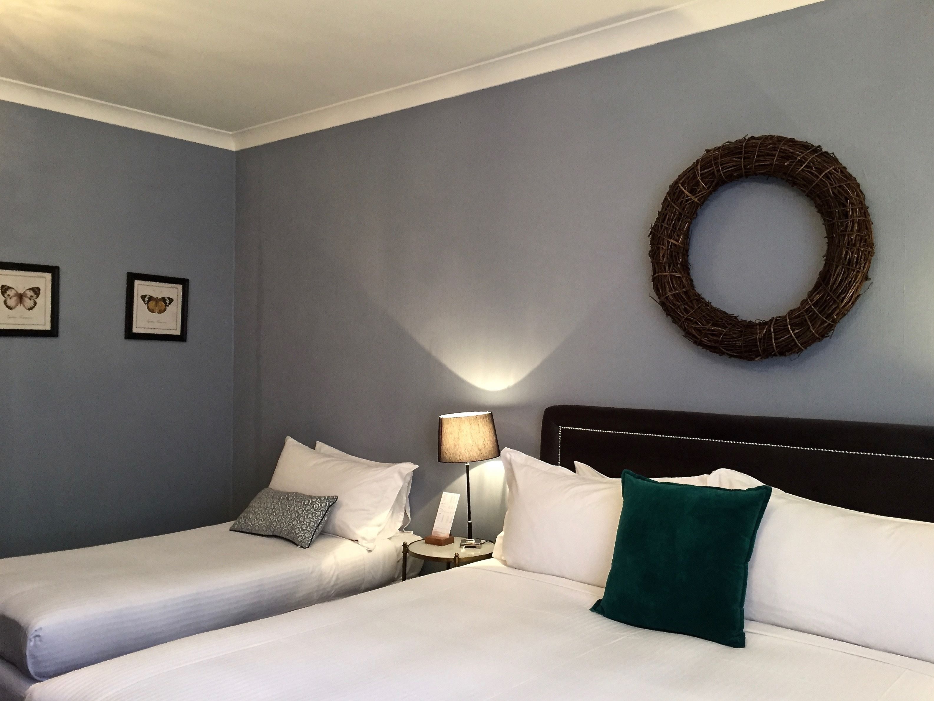 Habitación de hotel con dos camas individuales, una mesita de noche con lámpara, un arreglo en la pared y dos cuadros con mariposas.