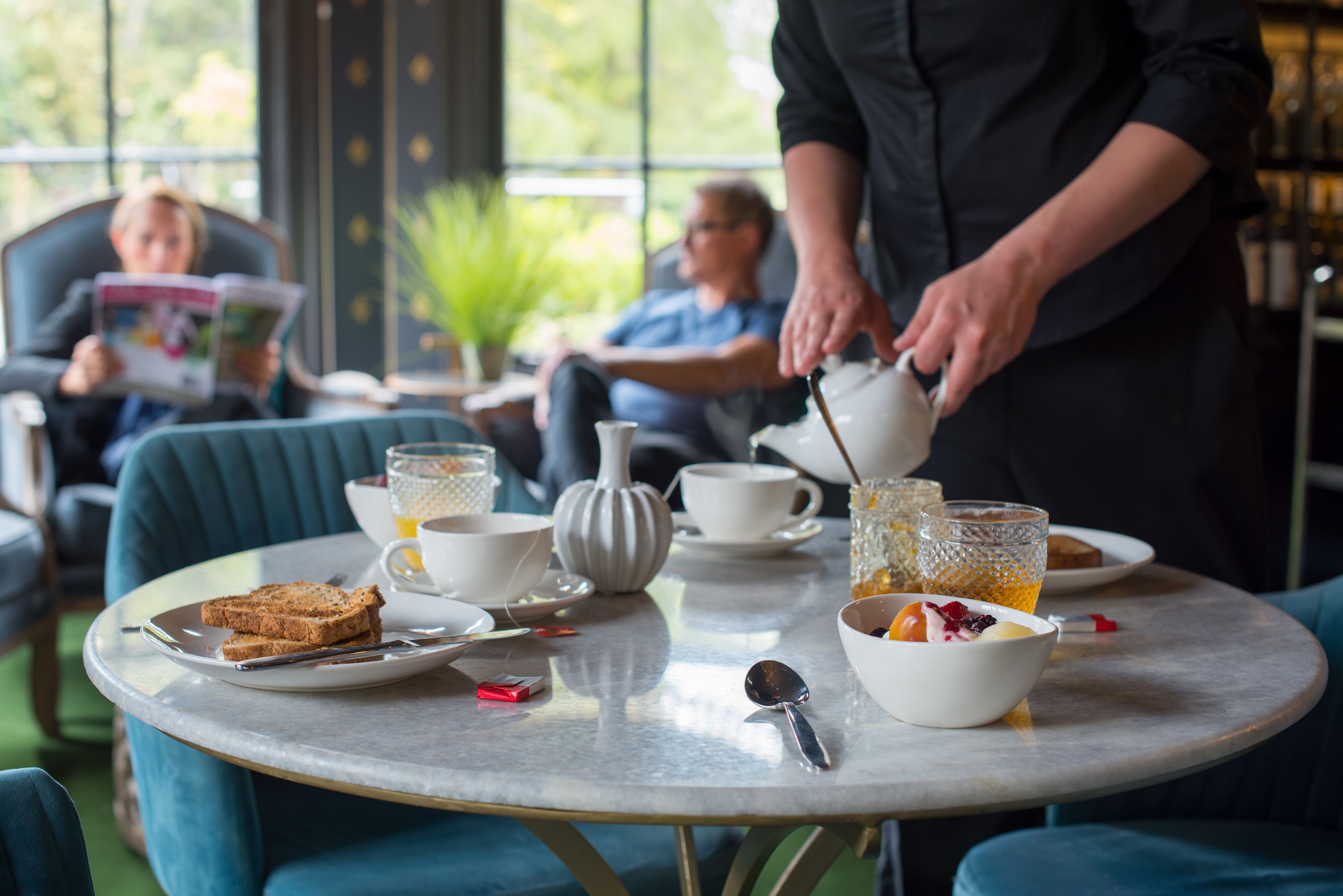A breakfast setting with a table set for two, including plates, cups, and a bowl of fruit.
