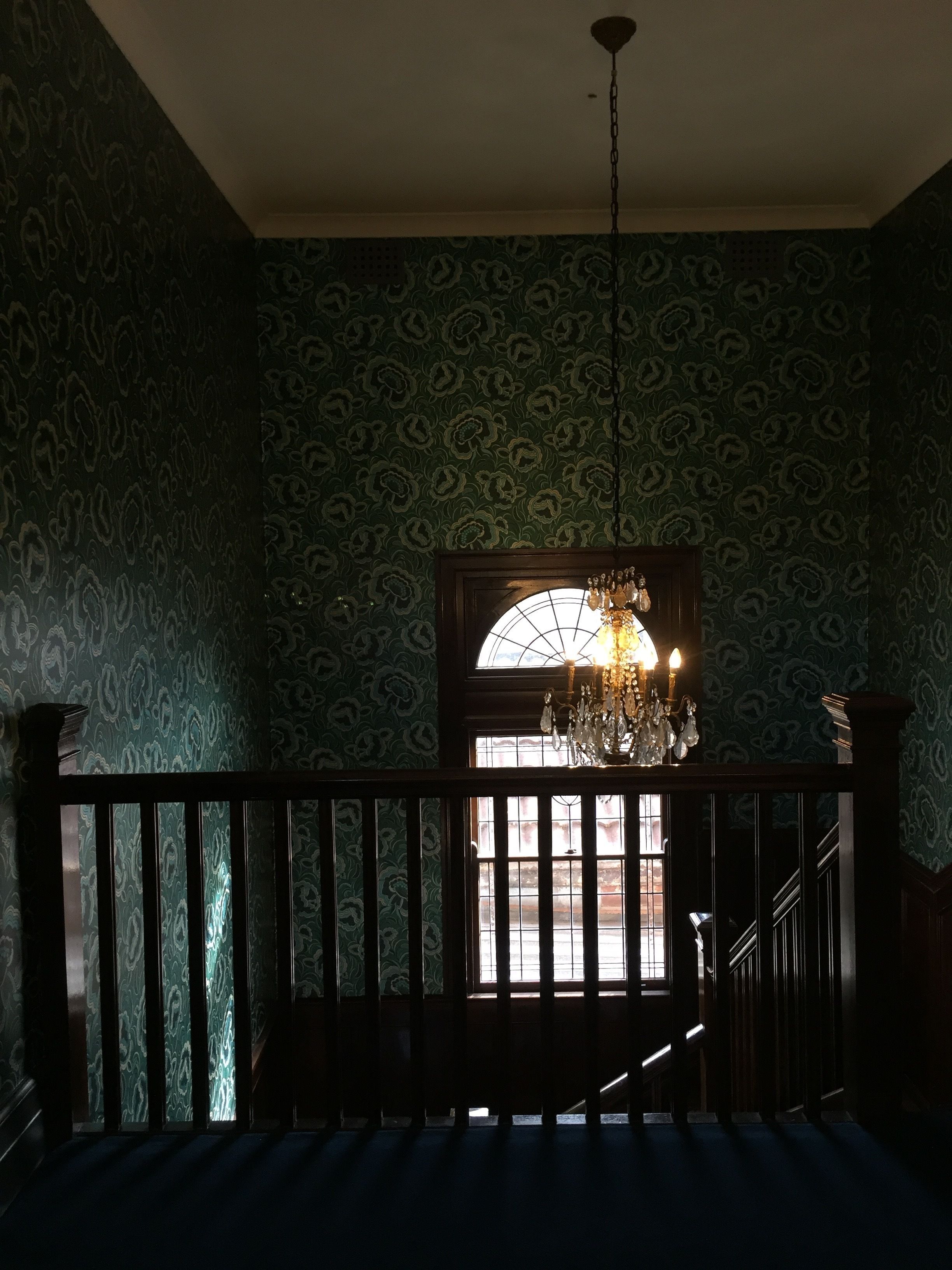 A room with patterned wallpaper, a chandelier, and a view through a window with a wooden railing.