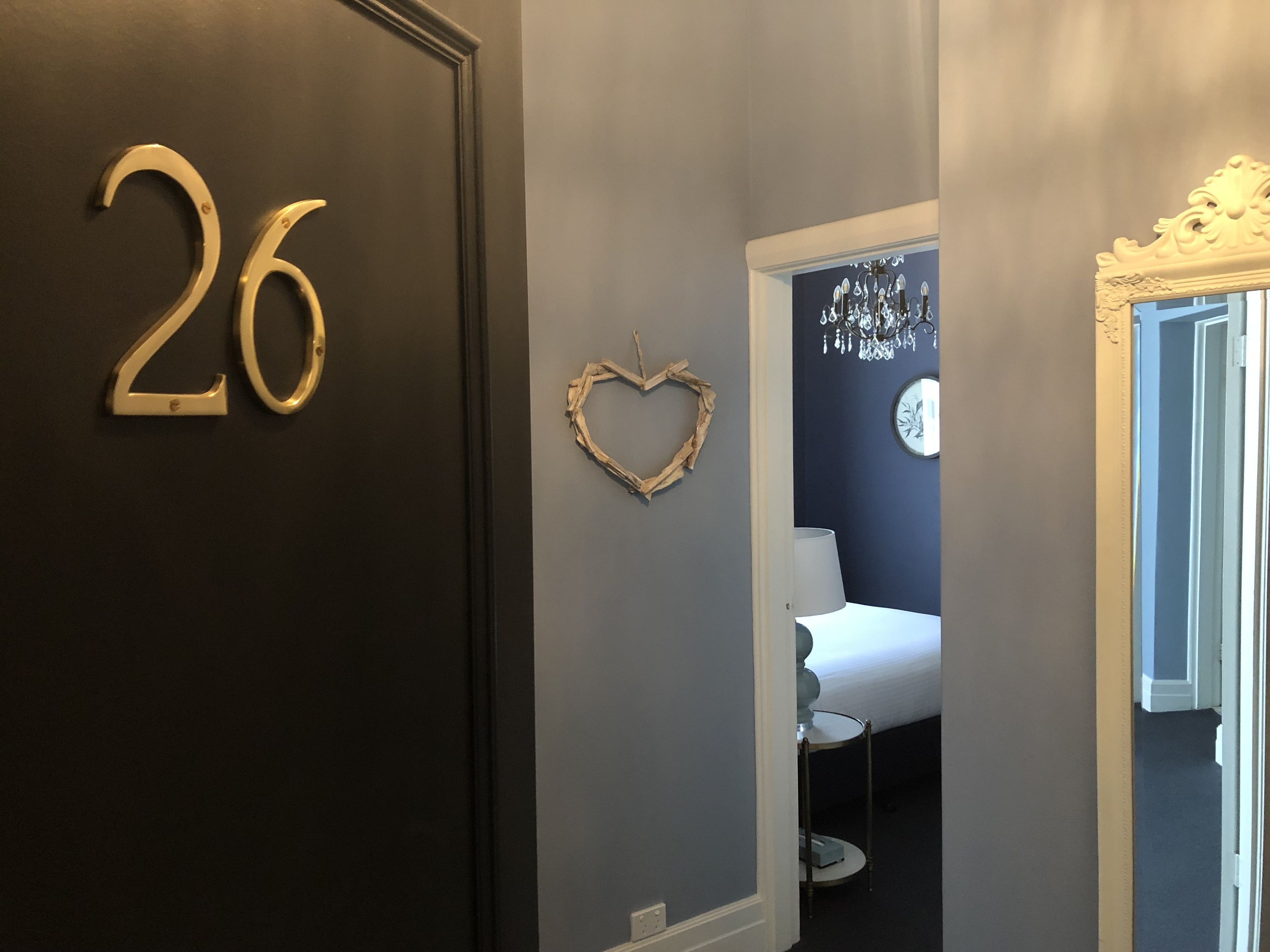 A room with a dark door marked with the number 26, a heart-shaped decoration on the wall, and a bed with a white headboard and a round mirror with an ornate frame.