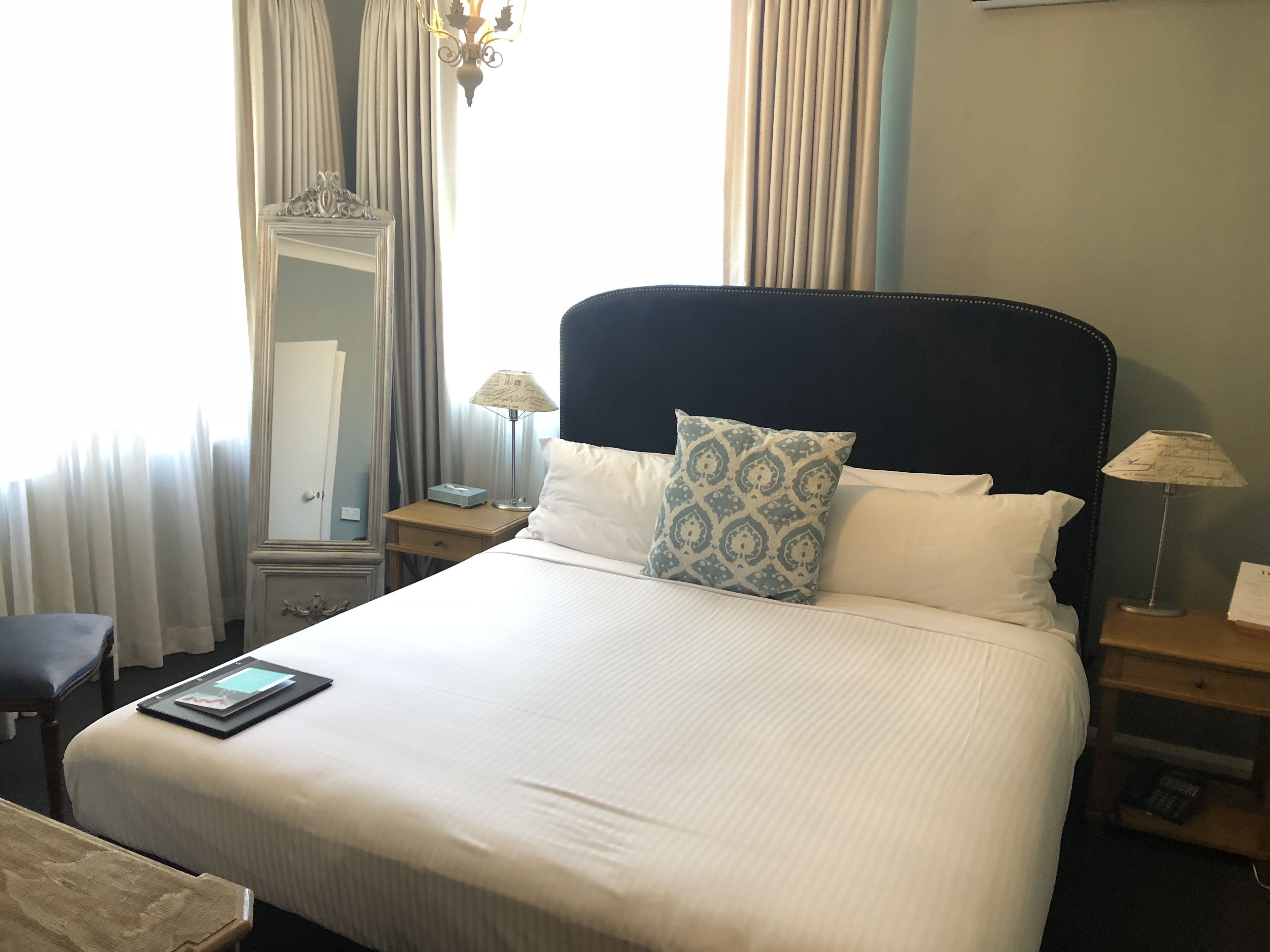 A neatly made bed with white linens and patterned pillows, a bedside table with a lamp, a mirror, and a chair.
