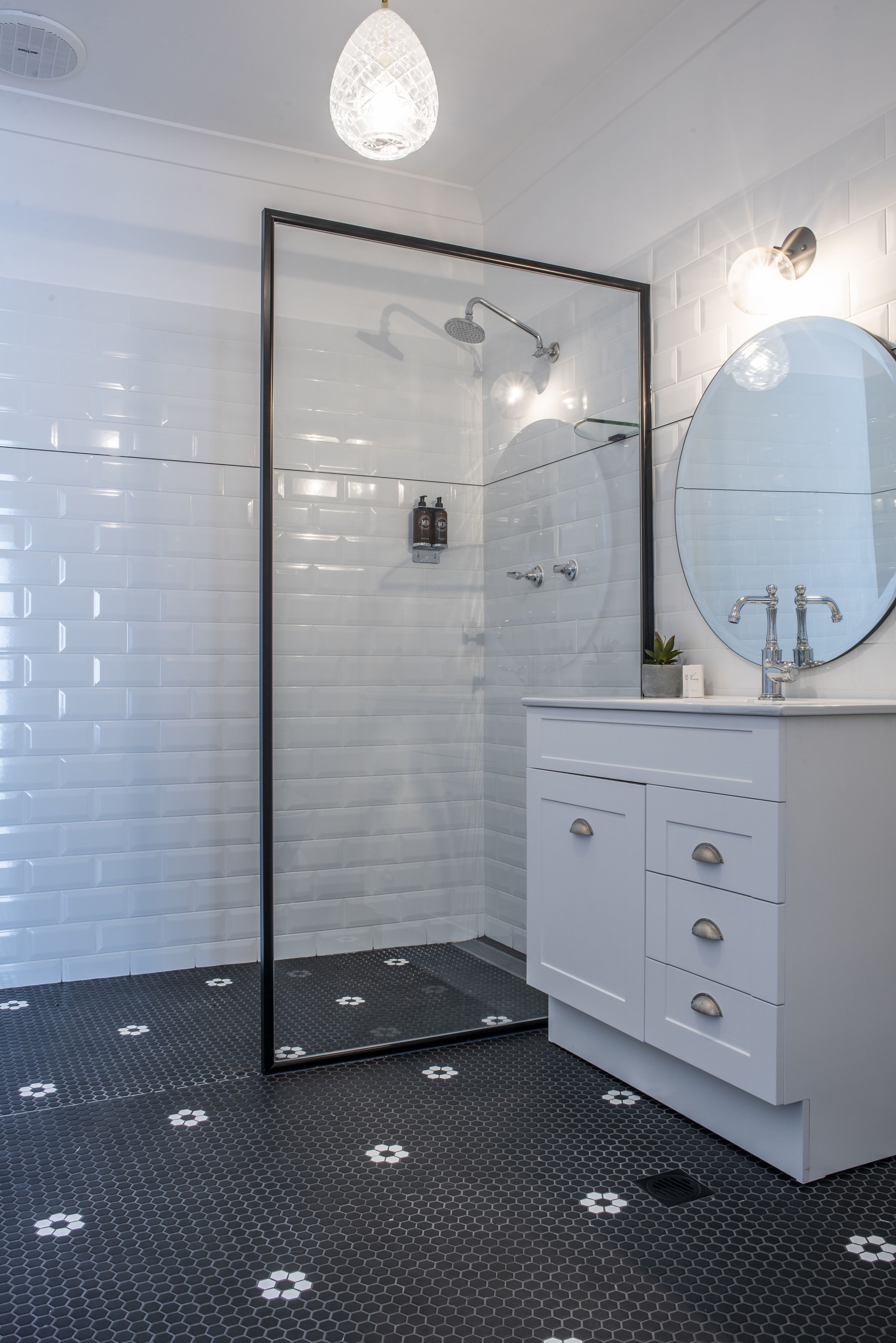 A modern bathroom with a glass shower enclosure, white tiled walls, a round mirror, and a white vanity with drawers.