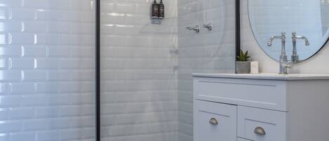 A modern bathroom with a glass shower enclosure, white tiled walls, a round mirror, and a white vanity with drawers.