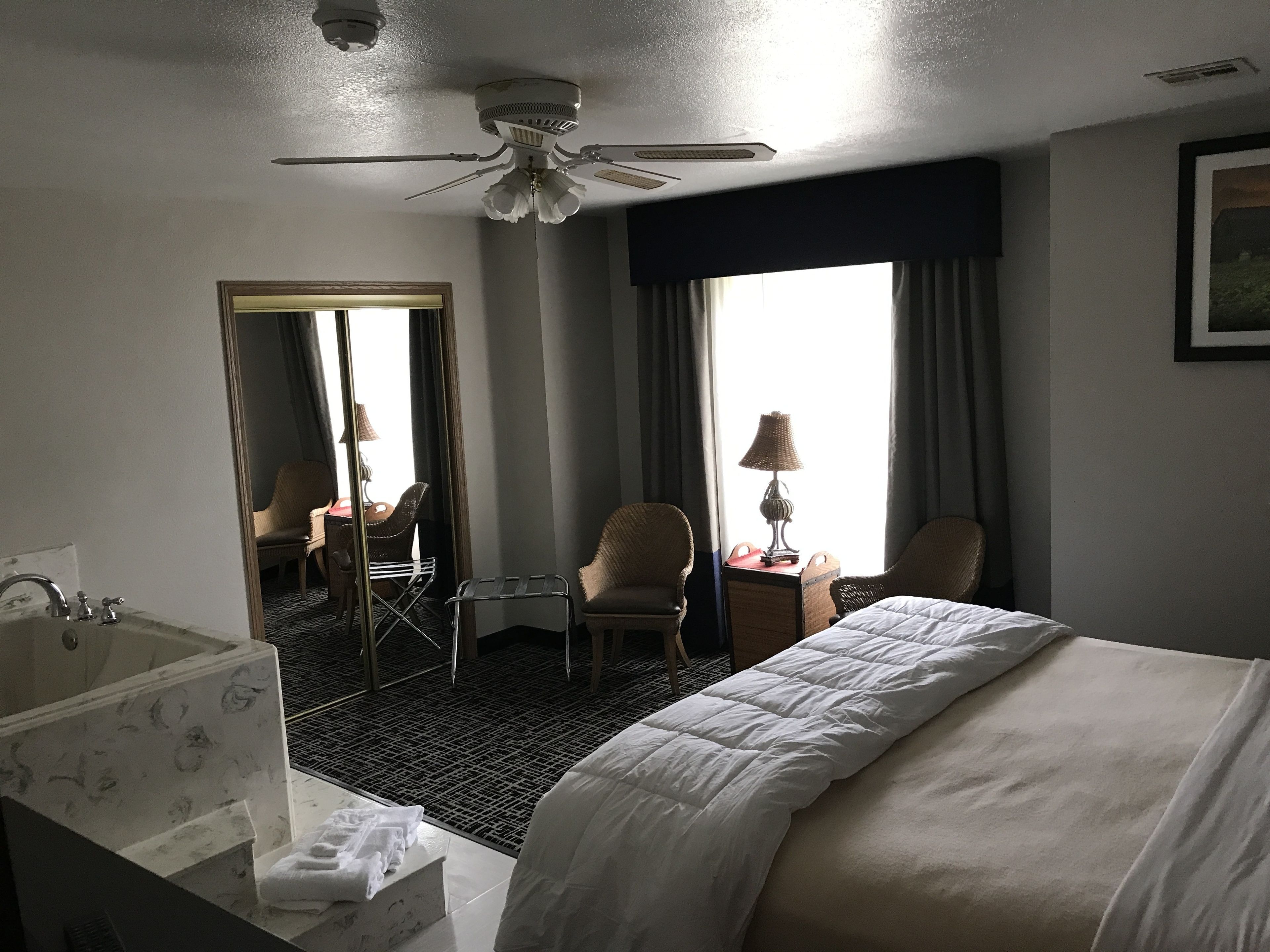 Habitación de hotel con cama, dos sillones, escritorio con lámpara, baño con bañera y ventilador de techo.