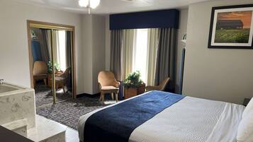 A hotel room with a bed, a chair, a desk, a mirror, a window with curtains, and a framed picture on the wall.