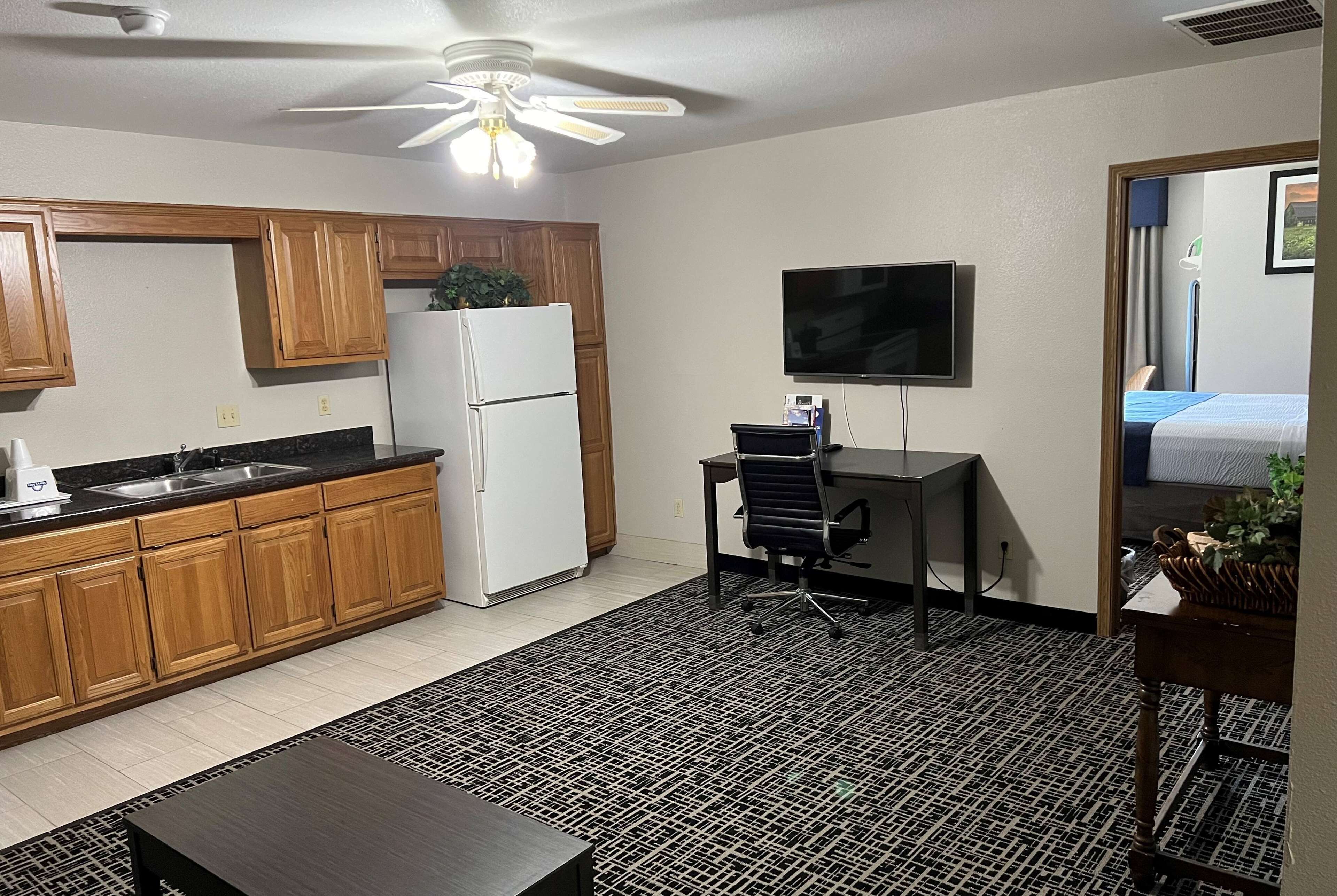 A compact kitchen with wooden cabinets, a refrigerator, a sink, and a ceiling fan. There's a small office area with a desk and chair, and a TV mounted on the wall.