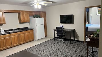 A compact kitchen with wooden cabinets, a refrigerator, a sink, and a ceiling fan. There's a small office area with a desk and chair, and a TV mounted on the wall.