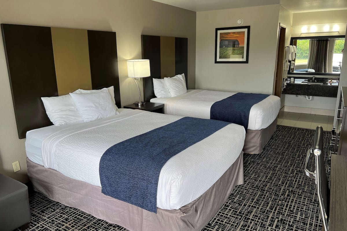 A hotel room with two beds, a nightstand with a lamp, a bathroom with a sink and mirror, and a patterned carpet.