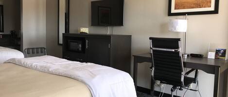 Habitación de hotel con una cama, un escritorio, una silla, un televisor, un microondas y un cuadro en la pared.