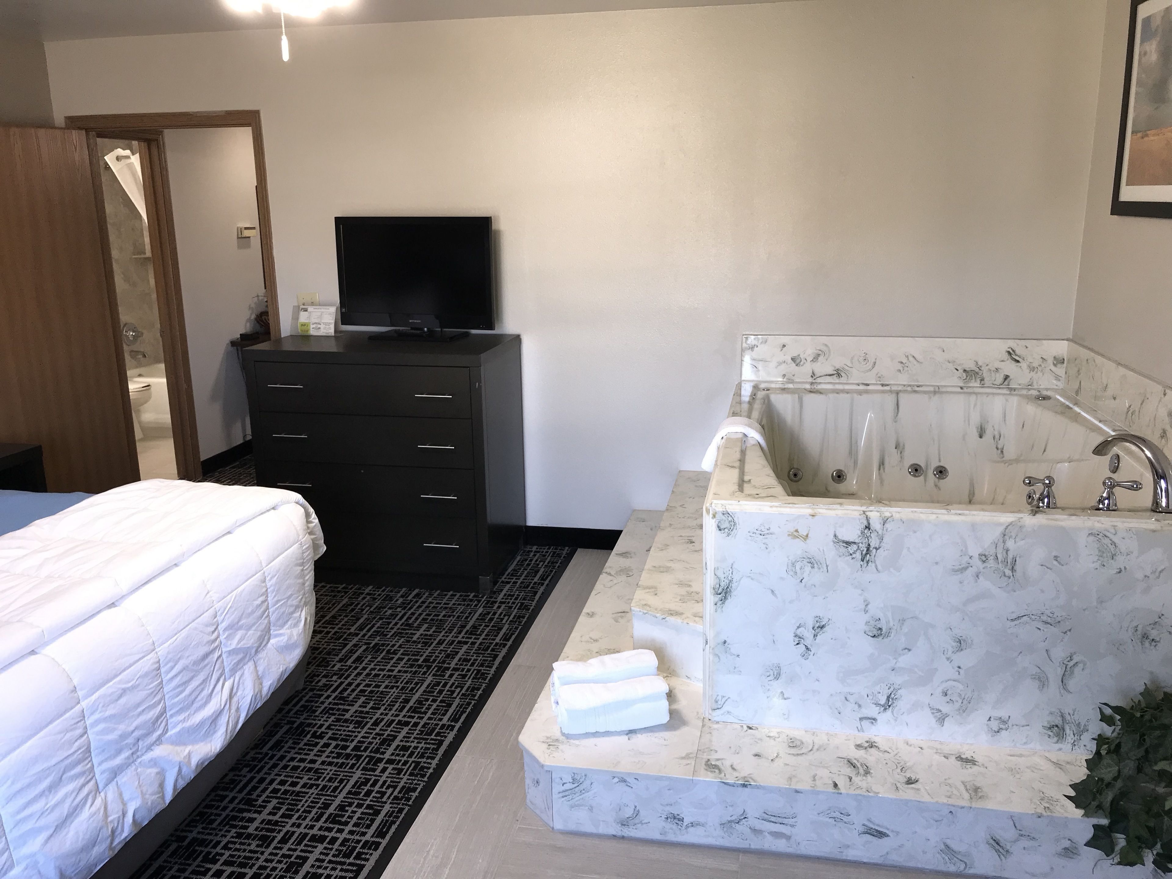 A hotel room with a bed, a black dresser, a television, and a marble-patterned bathtub with a shower.