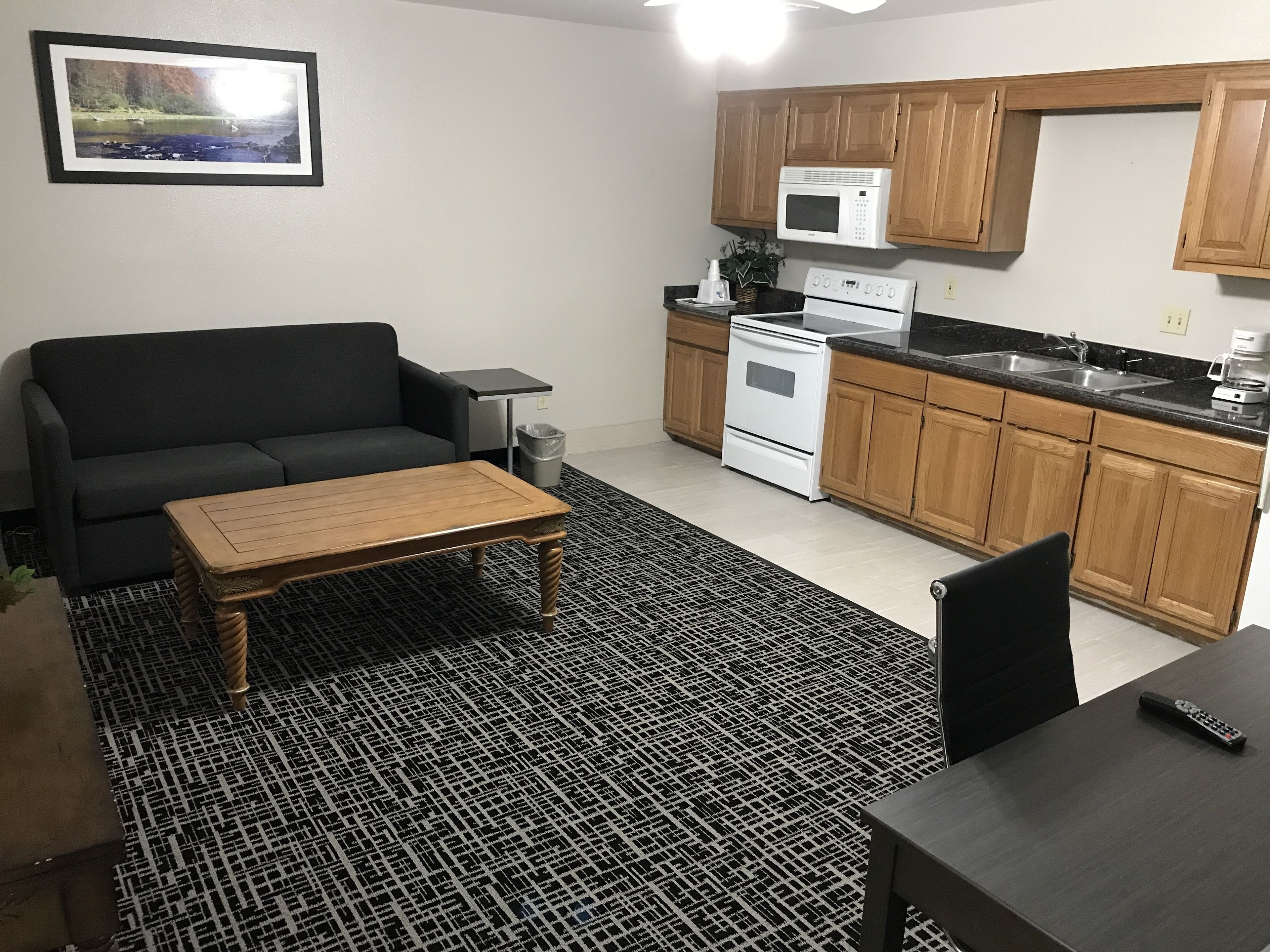 A compact living space with a sofa, a coffee table, a dining table, a kitchen with wooden cabinets and a microwave, and a patterned rug.