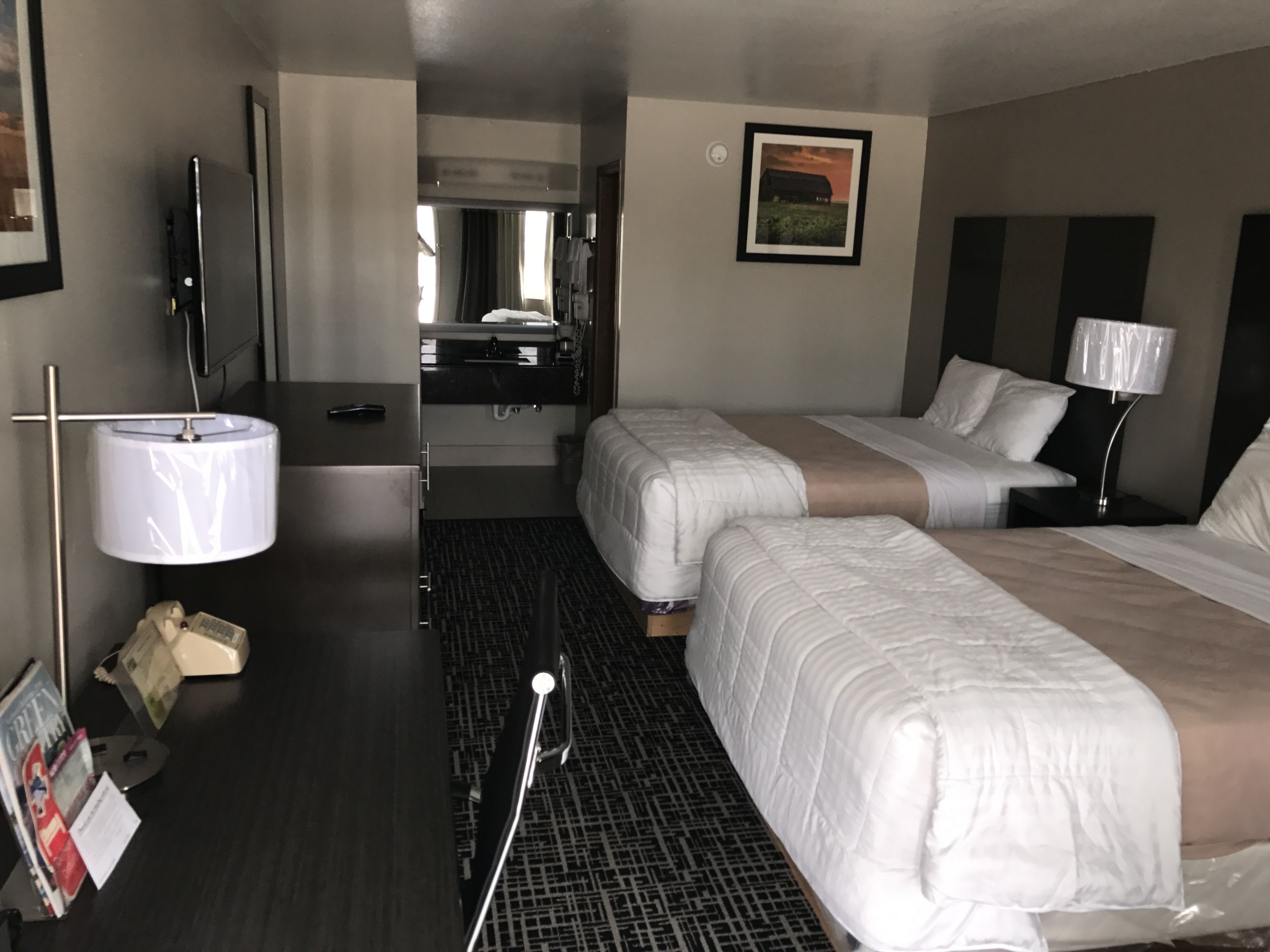A hotel room with two beds, a desk with a lamp, a mirror, a picture on the wall, and a small table with a magazine.