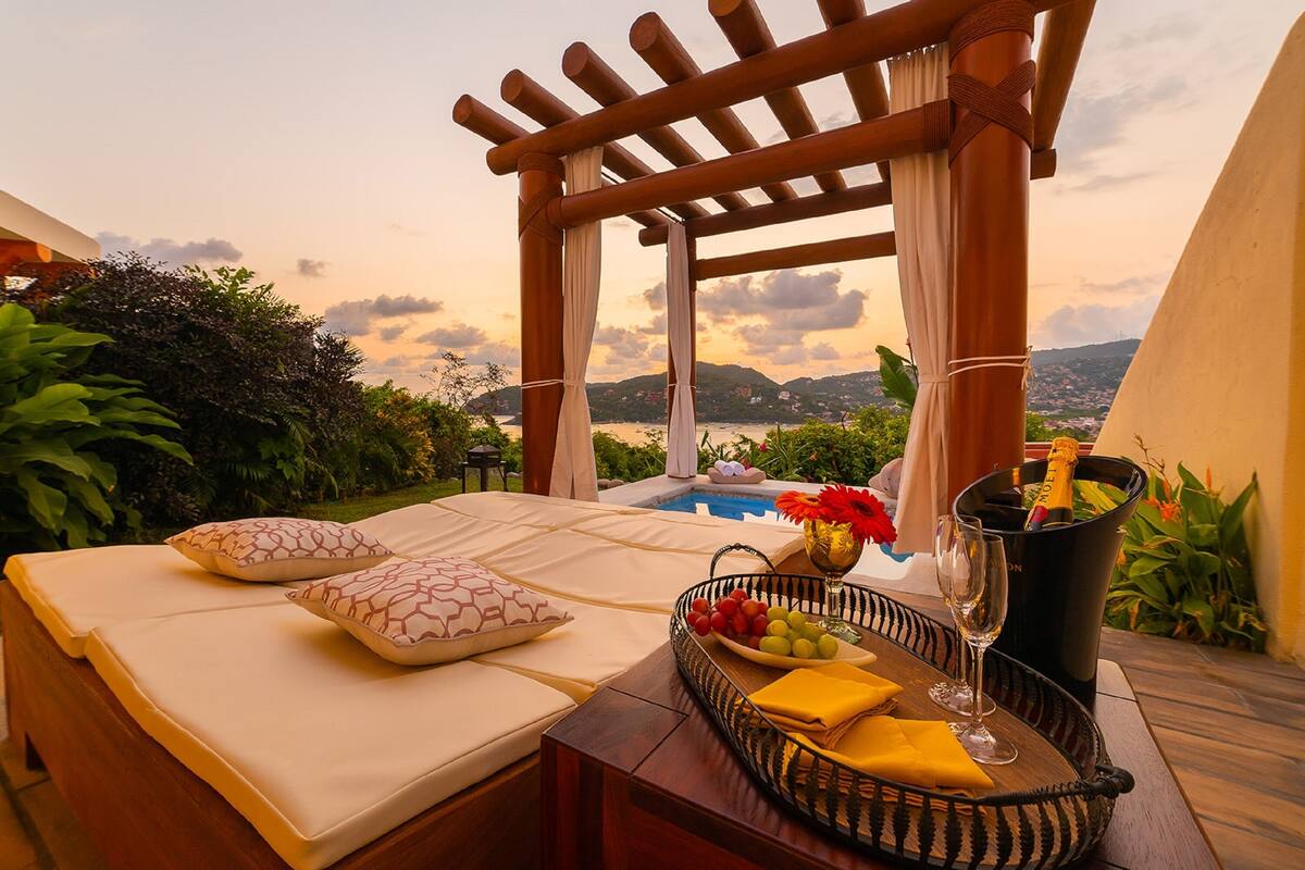 Un espacio al aire libre iluminado por el sol con piscina, una pérgola de madera y una mesa dispuesta con frutas, una botella de champán y copas.