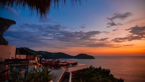 Blick vom Infinity-Pool eines Resorts auf das Meer bei Sonnenuntergang, mit einem Reetdach und einer Außenlounge.