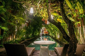 An indoor pool area with wicker lounge chairs and a central table, surrounded by lush tropical plants and a view of a building in the distance. - djabu Seminyak Hotel (Keroboken Kelod)