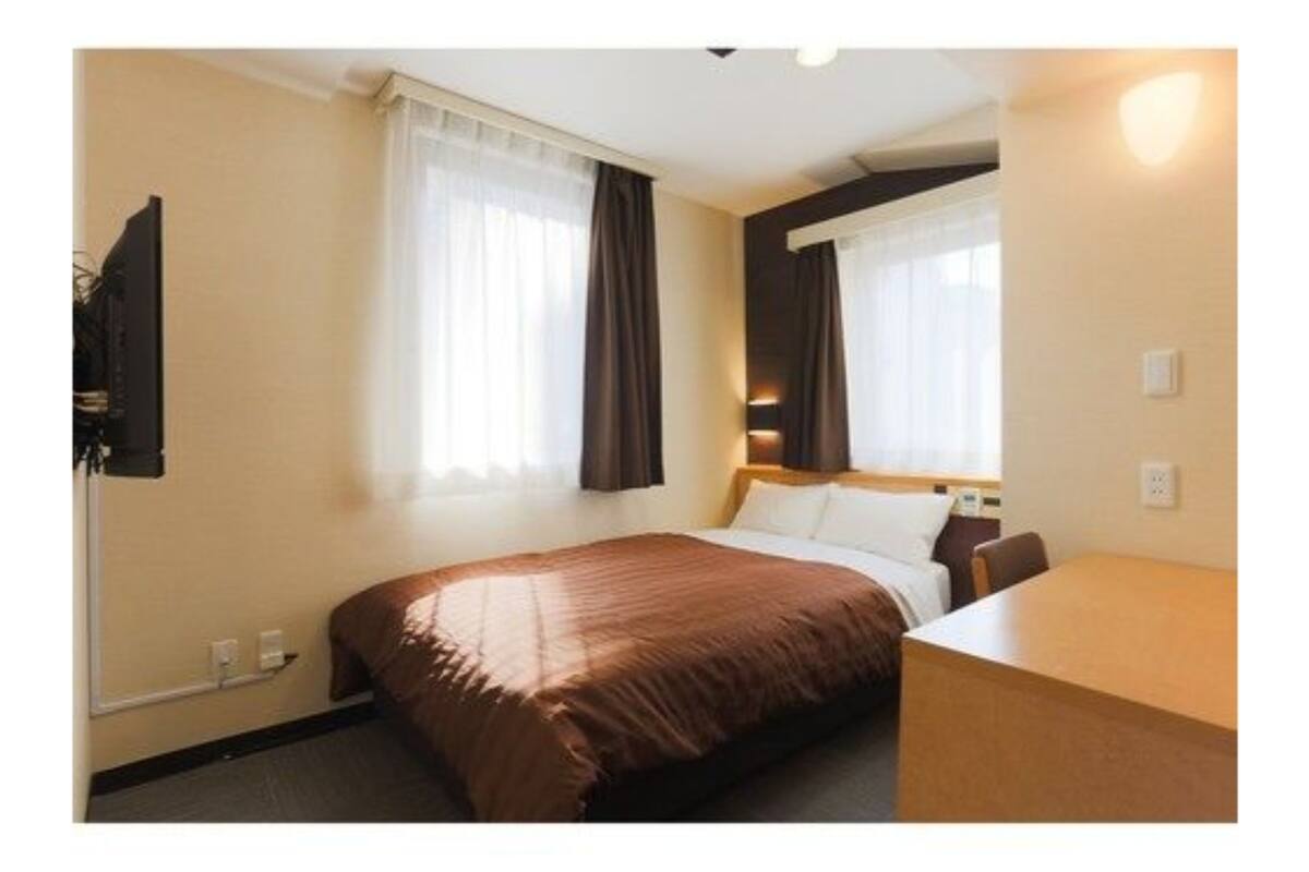 A hotel room with a bed, a desk, and two windows with curtains.