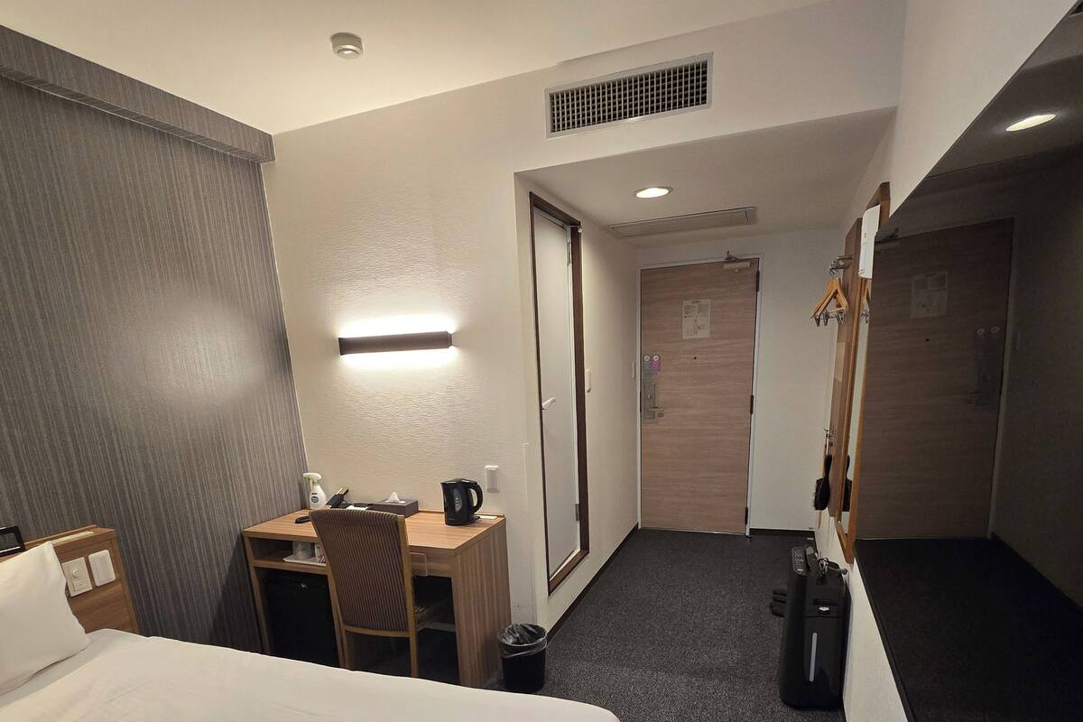 A hotel room with a bed, a desk, a chair, a coffee maker, a trash bin, and a coat rack.