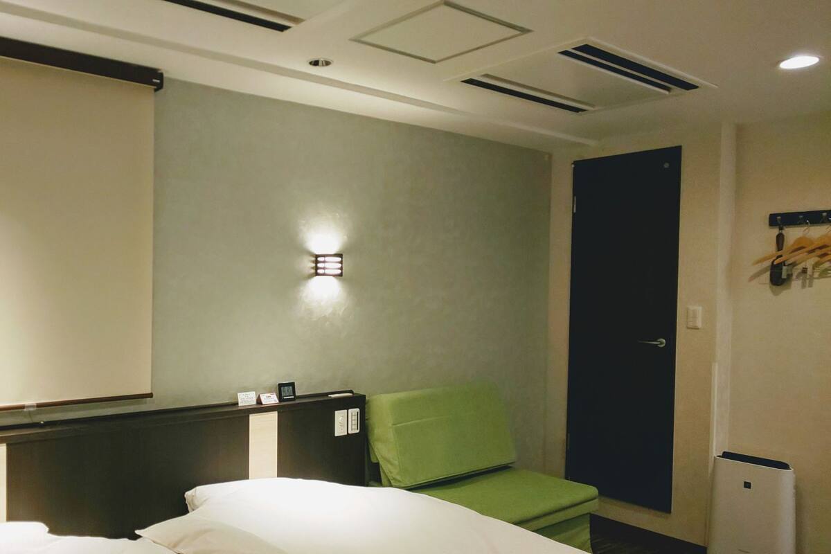 A hotel room with a bed, a green chair, a desk, and a wall-mounted light.